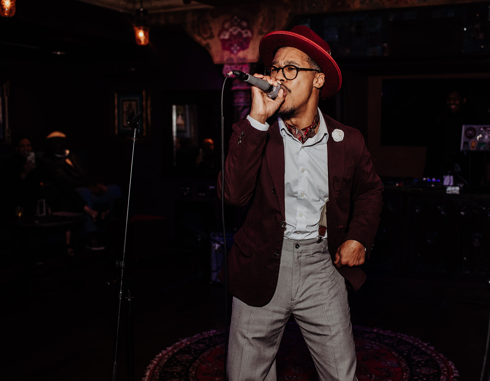 Person singing into microphone, wearing glasses, red hat, white shirt, dark blazer, and gray pants, in a dimly lit venue.