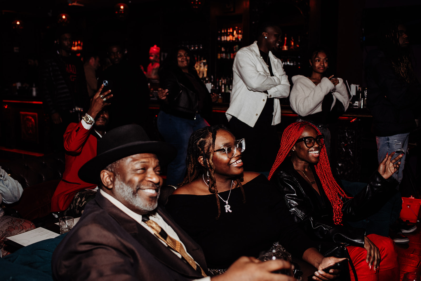 Group of people seated at a bar or lounge, some smiling and engaging, with a dark background and warm lighting.
