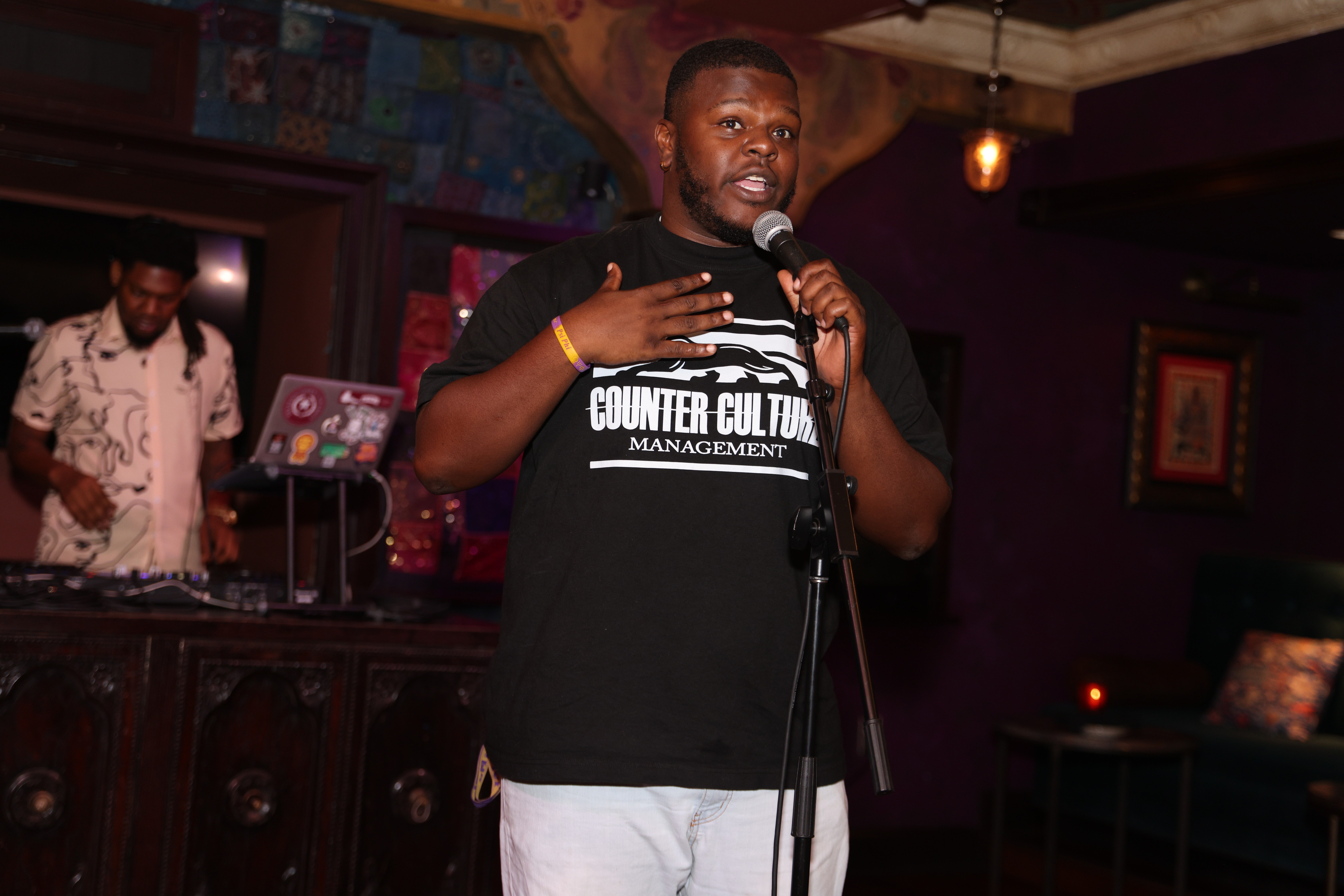 Man speaking into microphone on stage with DJ in background, holding hand on chest, in a dimly lit venue.