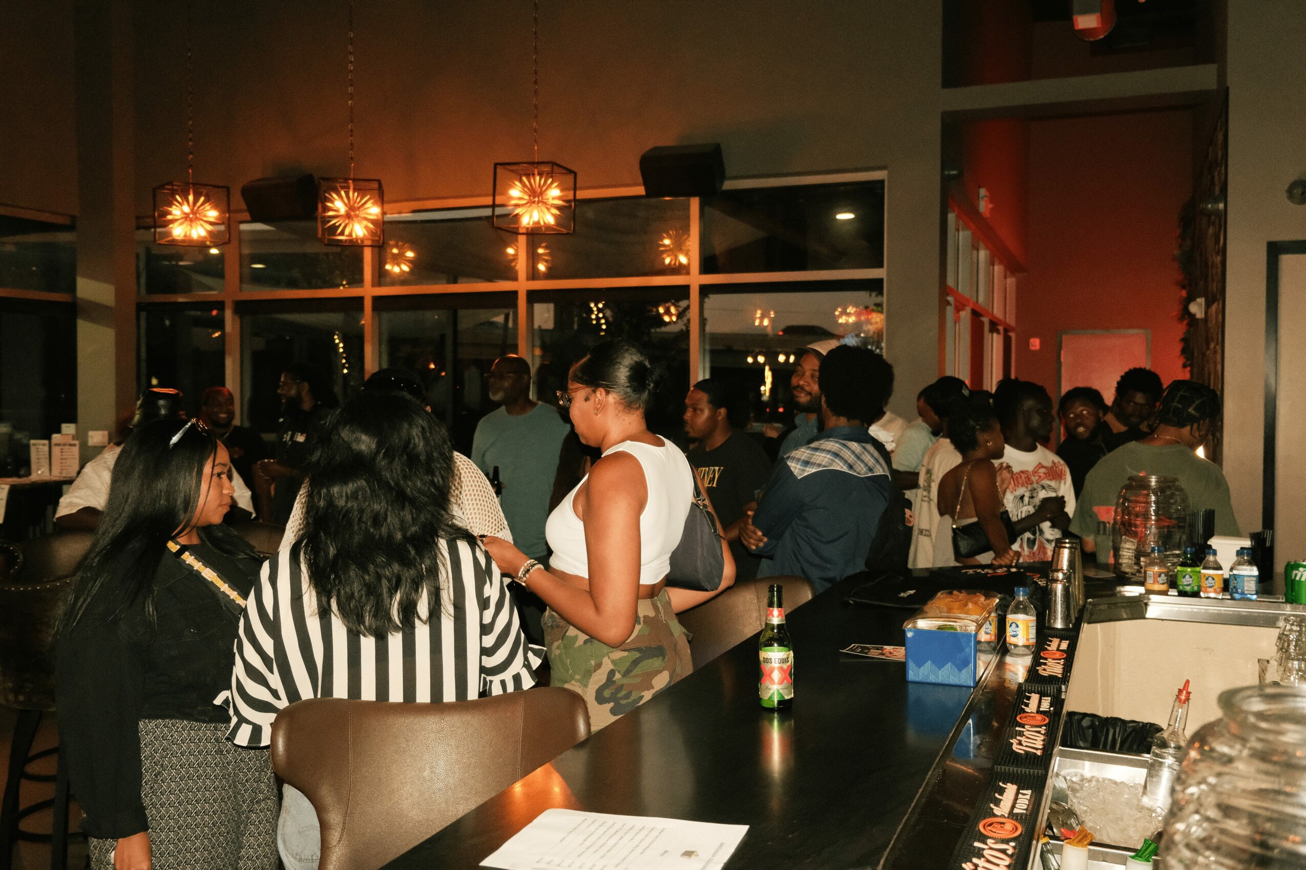 Crowded indoor space with people socializing, bar area with bottles, and warm lighting fixtures.