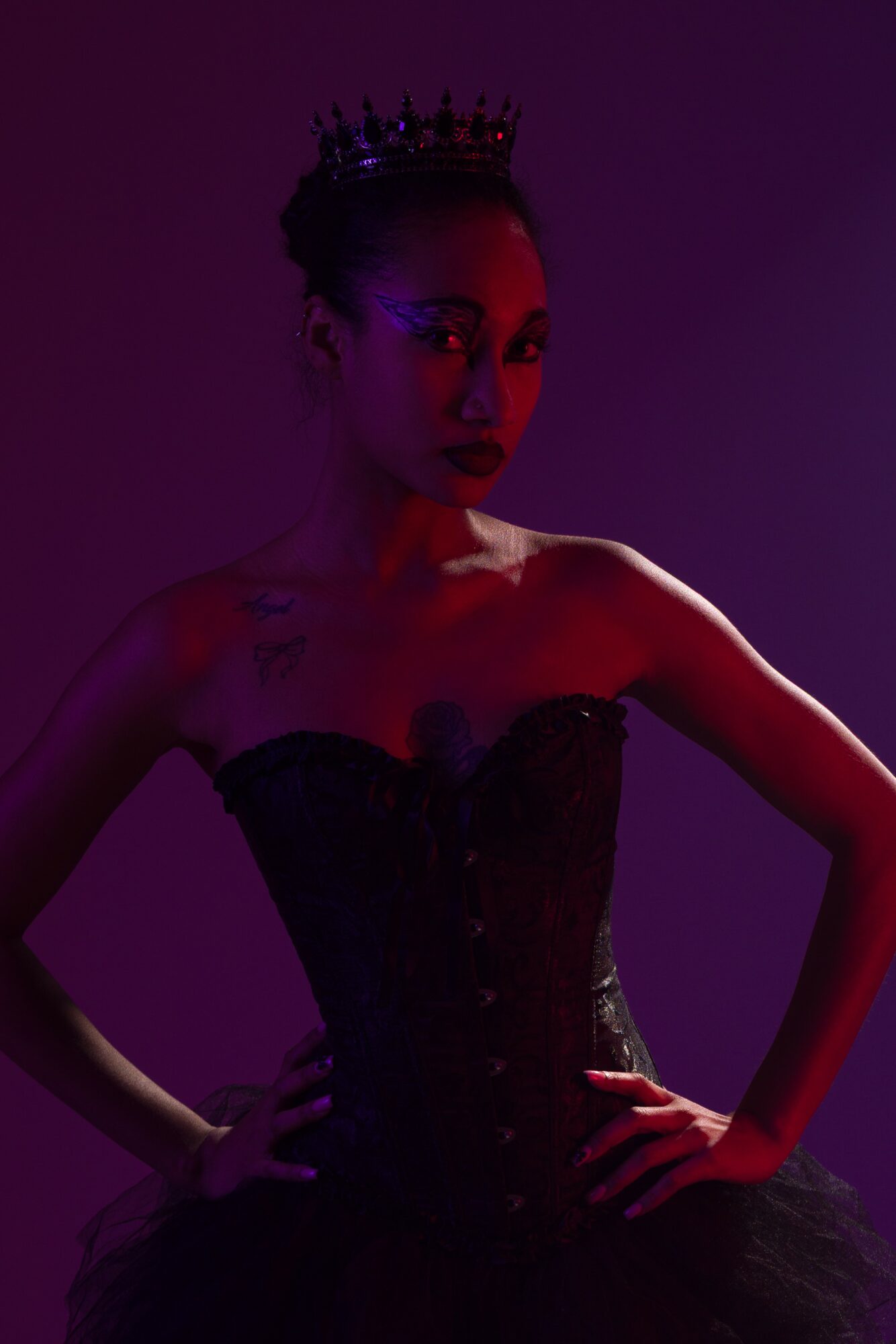Woman wearing a crown and strapless dress, posing with hands on hips, in dark lighting.