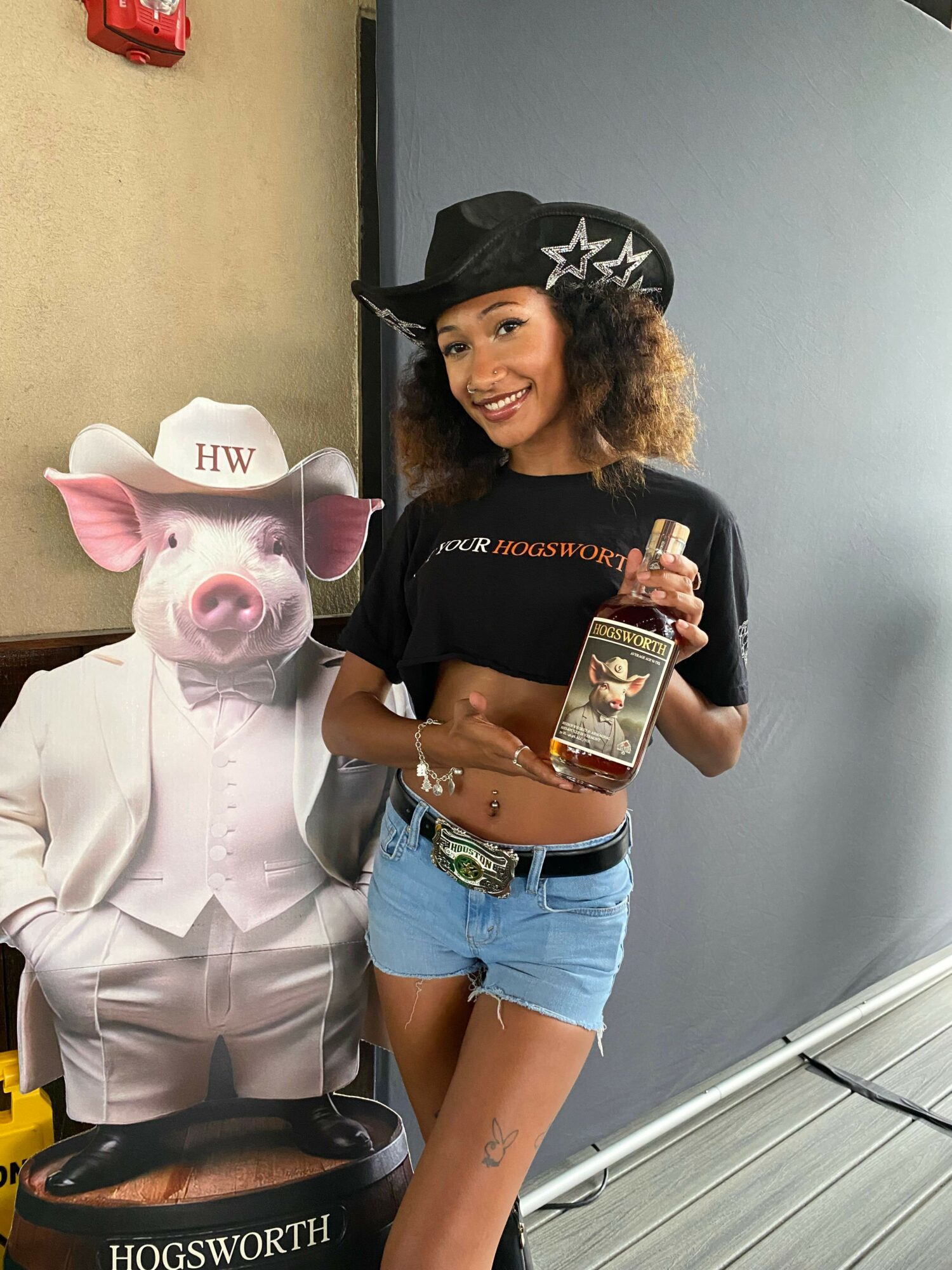 Young woman in a cowboy hat holding a phone, smiling, standing next to a person in a pig costume with a hat labeled 'HW'.