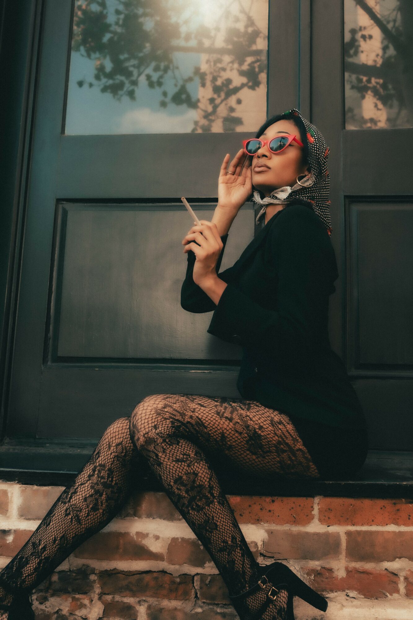 Woman sitting on brick ledge, wearing sunglasses and patterned tights, holding a cigarette, in front of a window with trees outside.