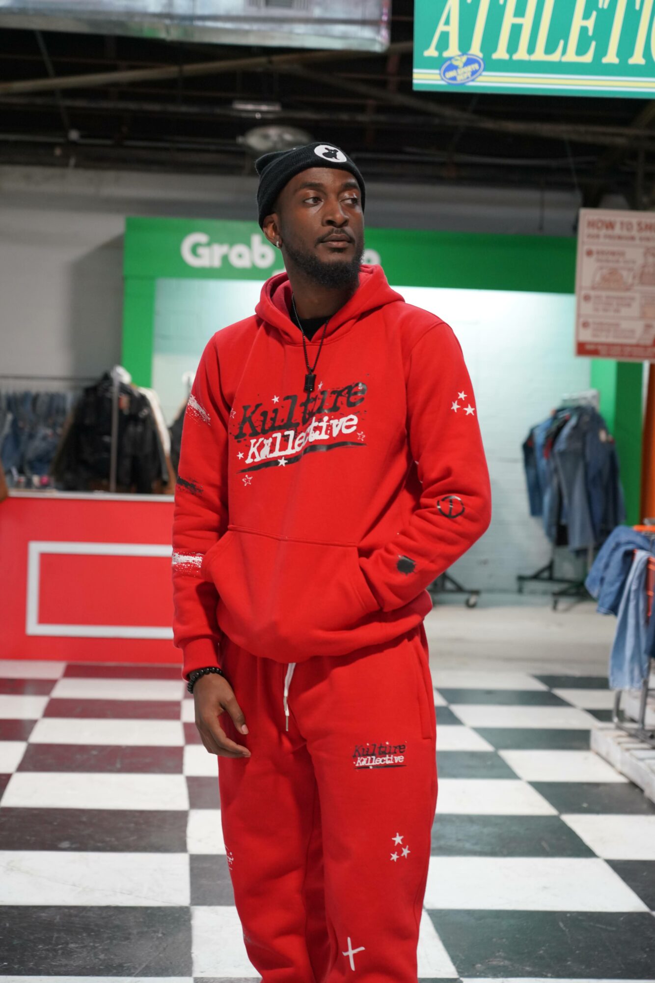 Young man in red hoodie and sweatpants standing in store with checkered floor and clothing racks.