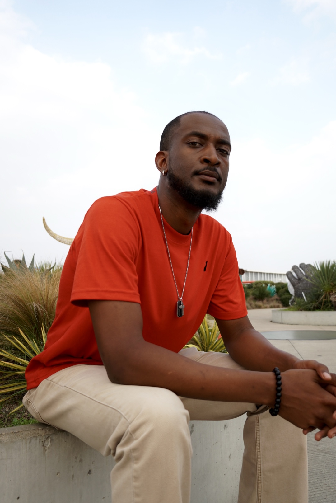 Man sitting outdoors on a concrete ledge, wearing a red shirt and beige pants, with a necklace and bracelet.