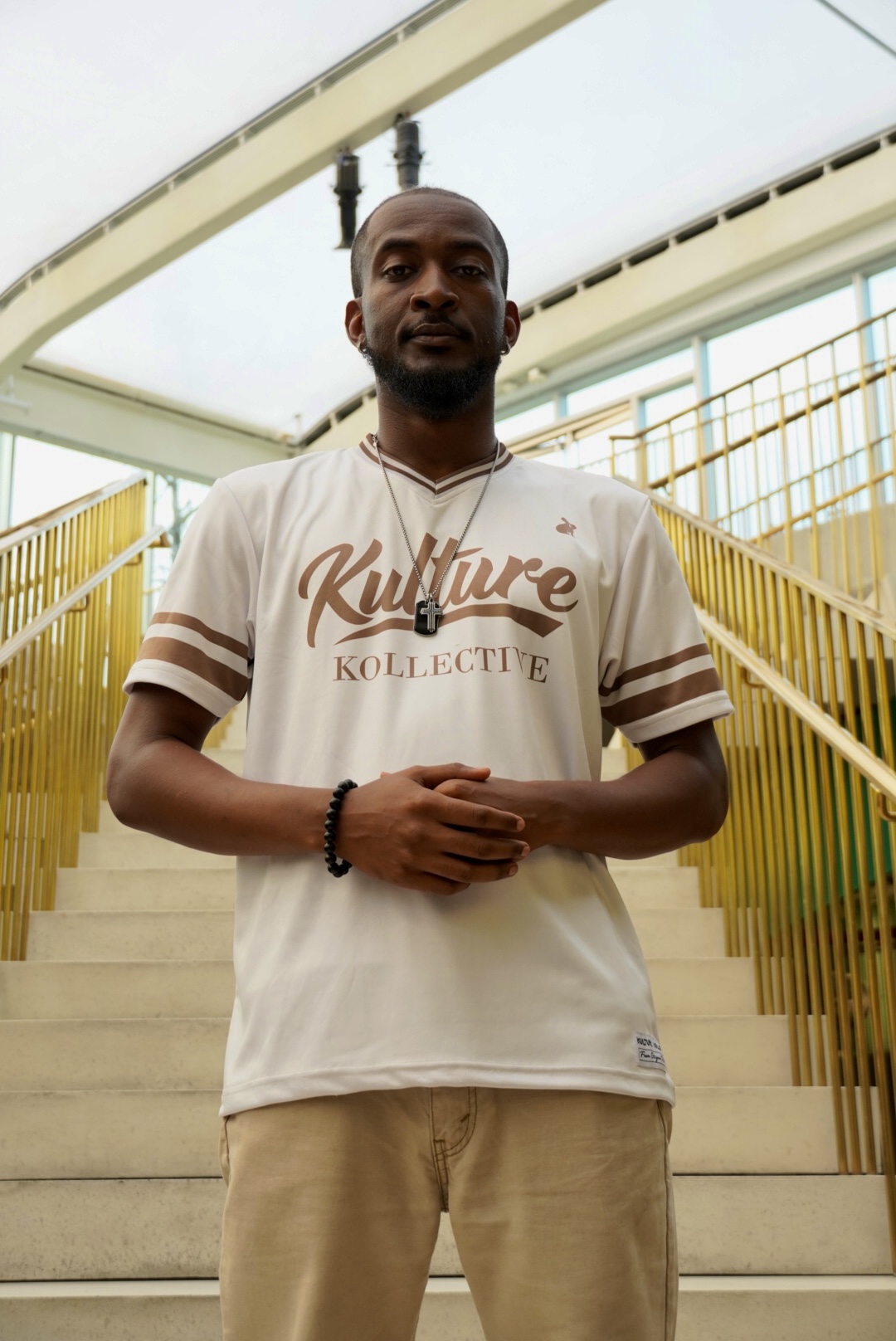 Man standing on stairs with yellow railings, wearing a white T-shirt and beige pants, in an outdoor setting.