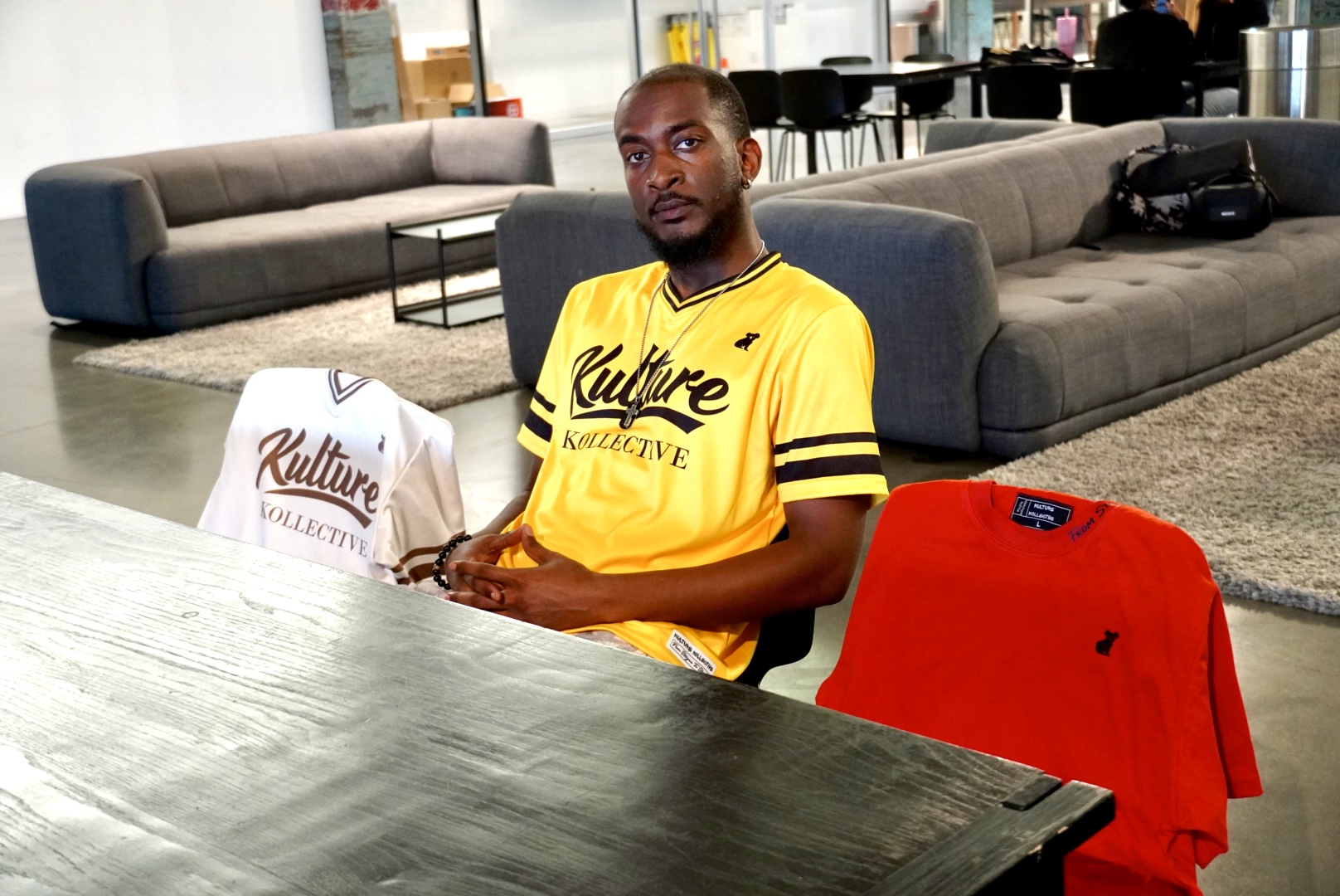 Man in yellow sports jersey sitting at a table in a modern lounge area with sofas and large windows.