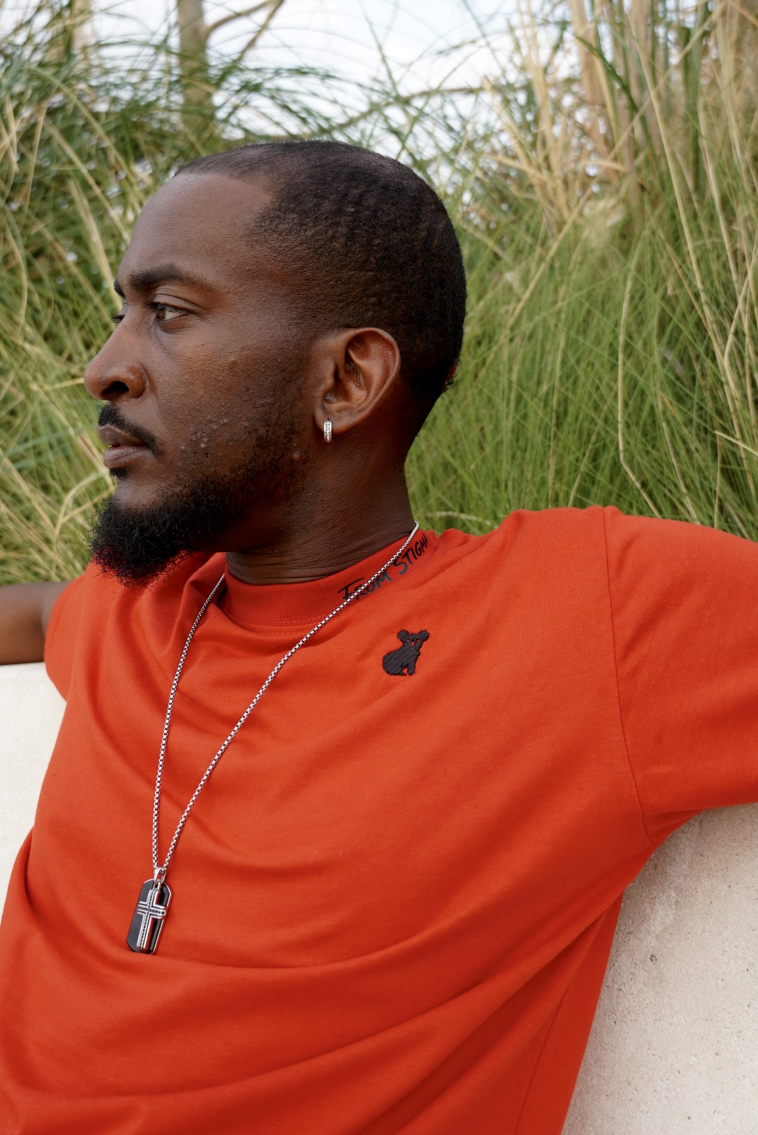 Man with short hair and beard wearing a red shirt, sitting outdoors near tall grass.