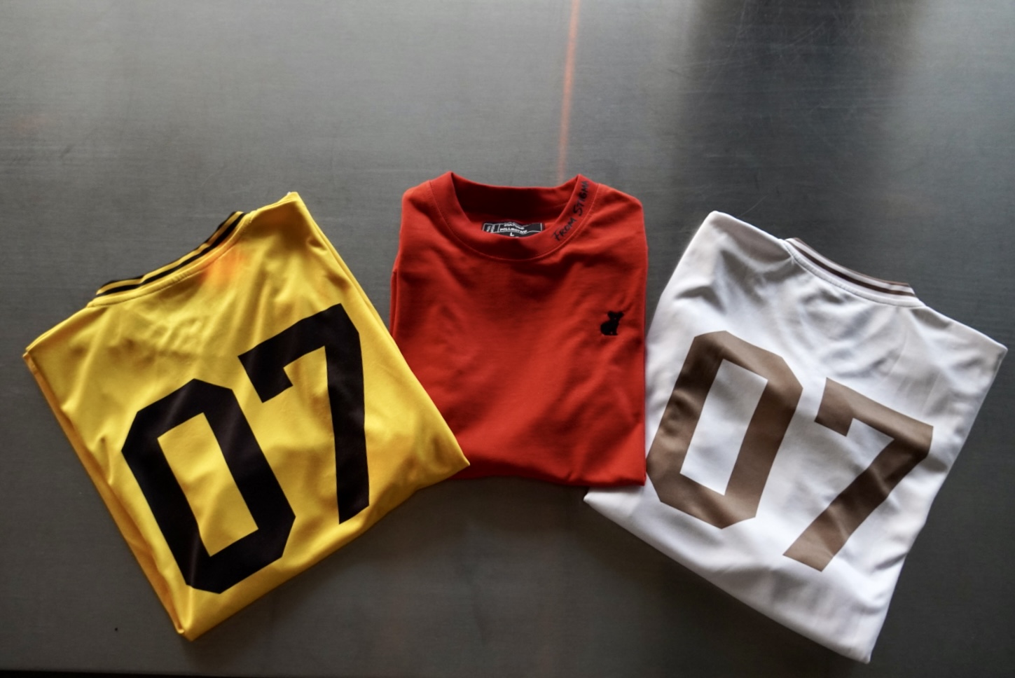 Three folded shirts on a dark surface, yellow with black number 07, red, and white with brown number 07.