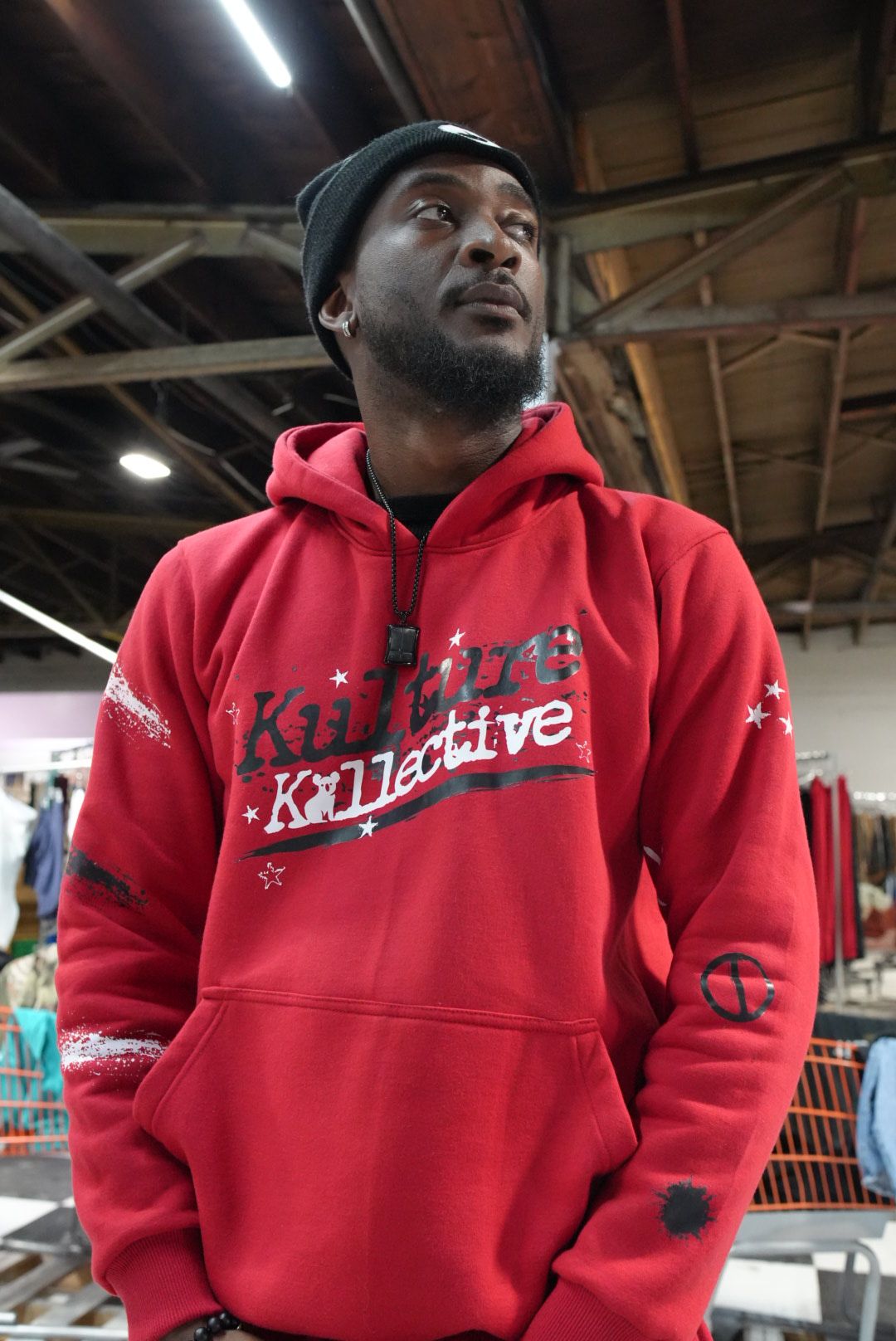 Man wearing a black beanie and red hoodie with text, standing indoors with ceiling beams visible.