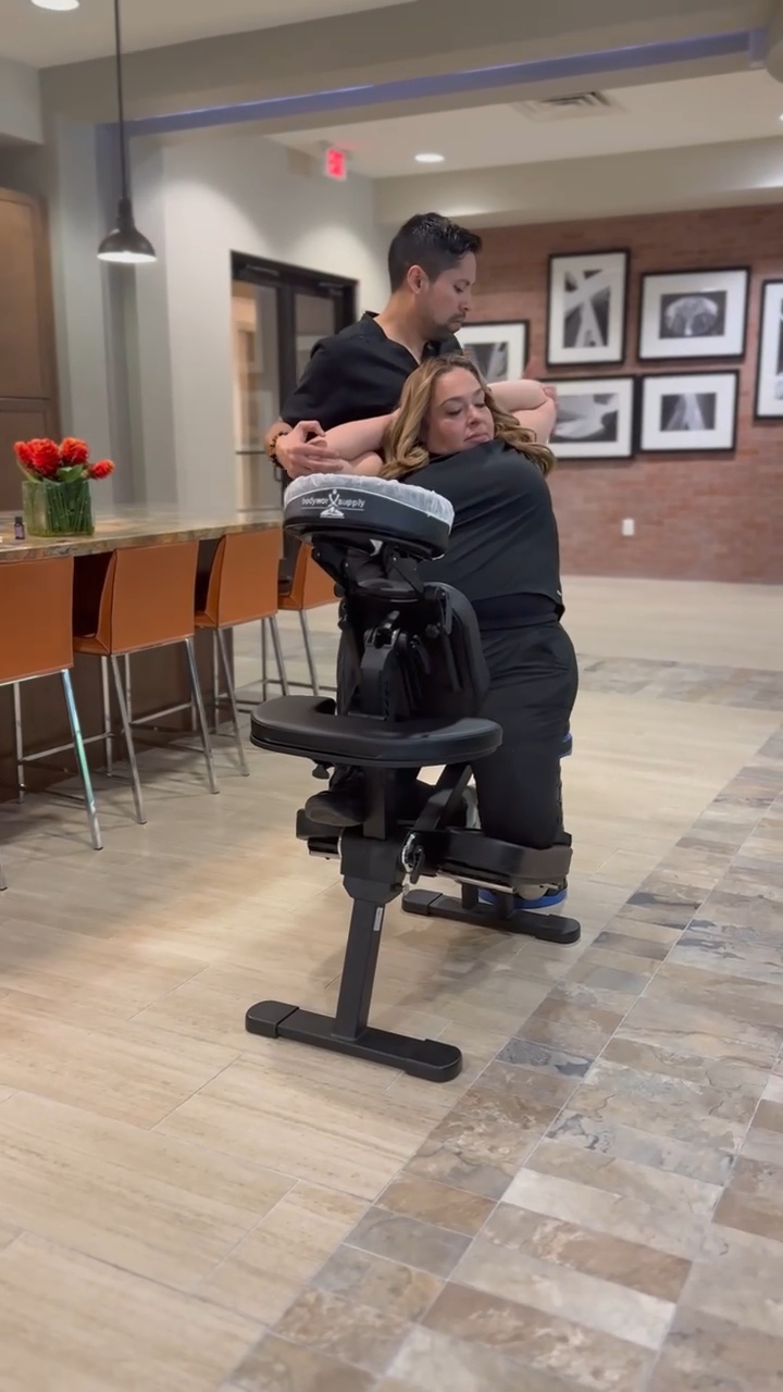 A person receiving a massage from a massage therapist in a room with framed pictures on the wall.