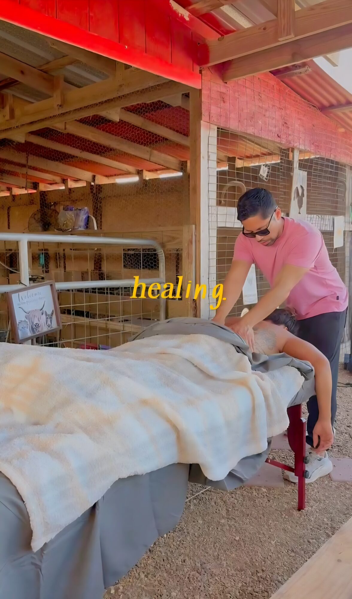 Person in pink shirt and glasses leaning over a bed with a blanket, in an outdoor setting with wooden structures.