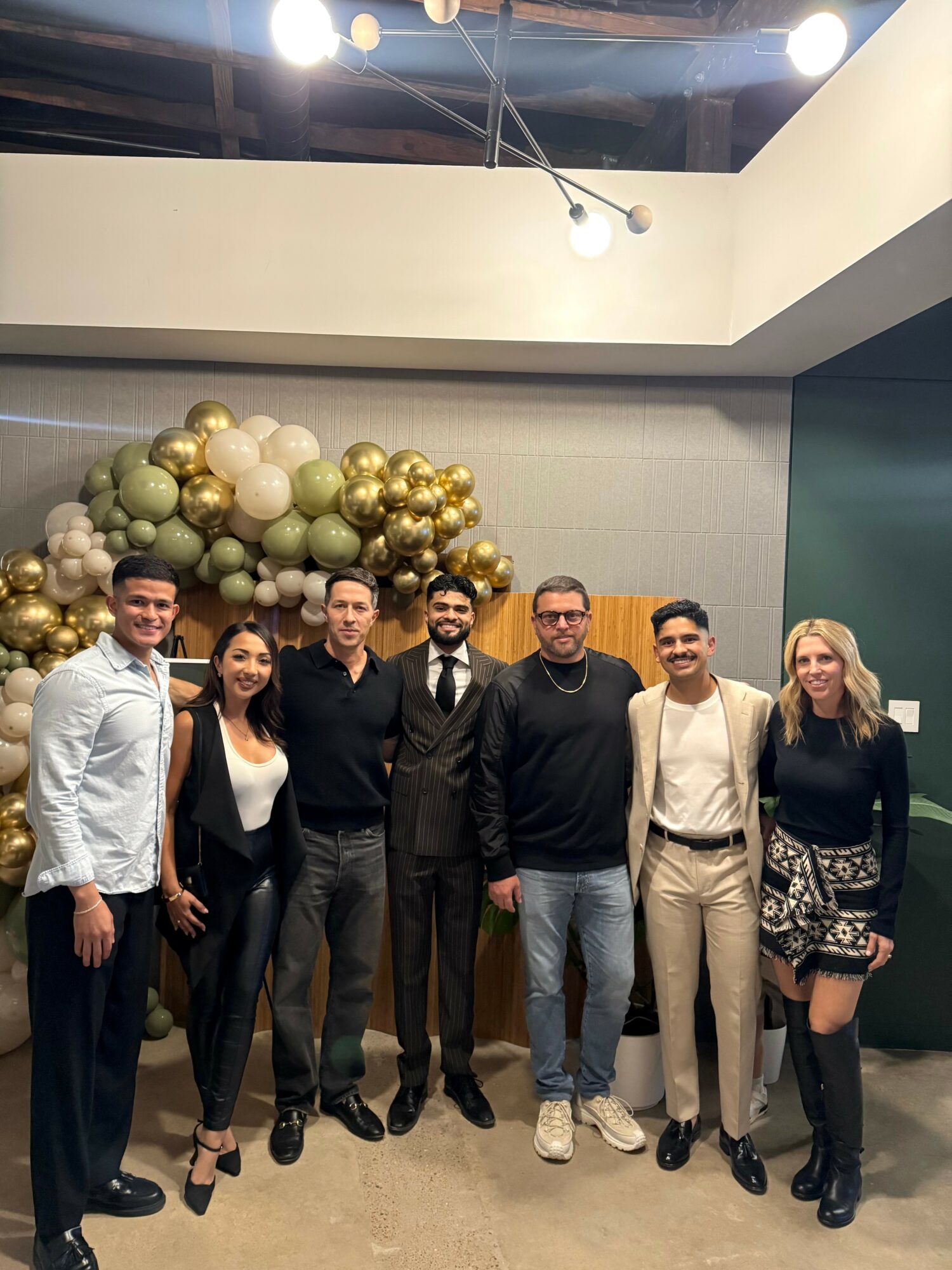 Group of seven people standing indoors with balloon decorations behind them, smiling at the camera.