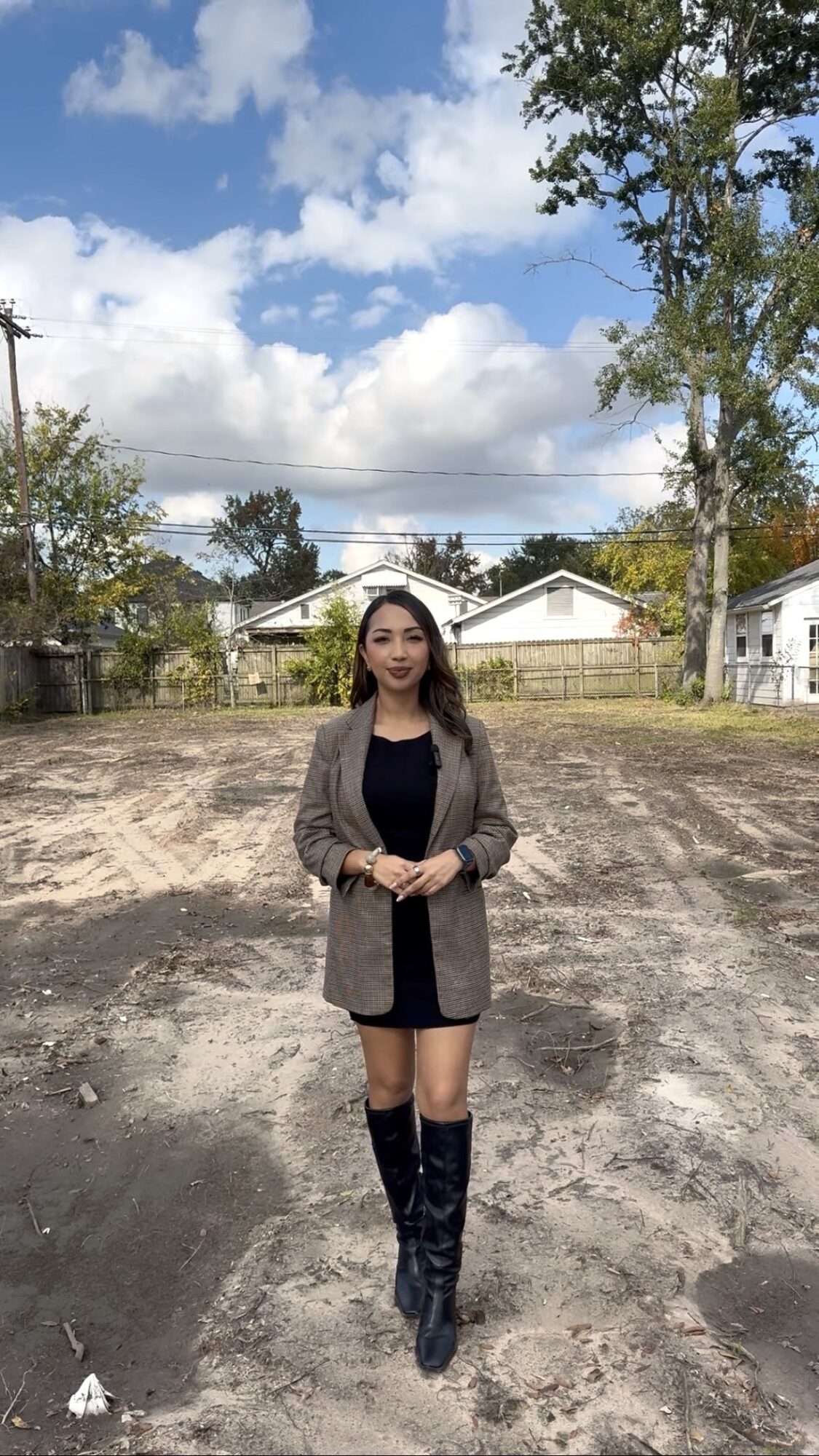 Woman standing outdoors on dirt ground with trees and houses in background.