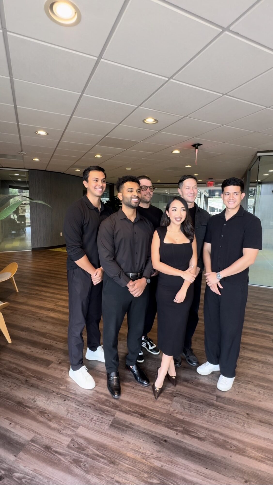 Group of six people standing indoors, smiling, dressed in black, with a modern interior background.