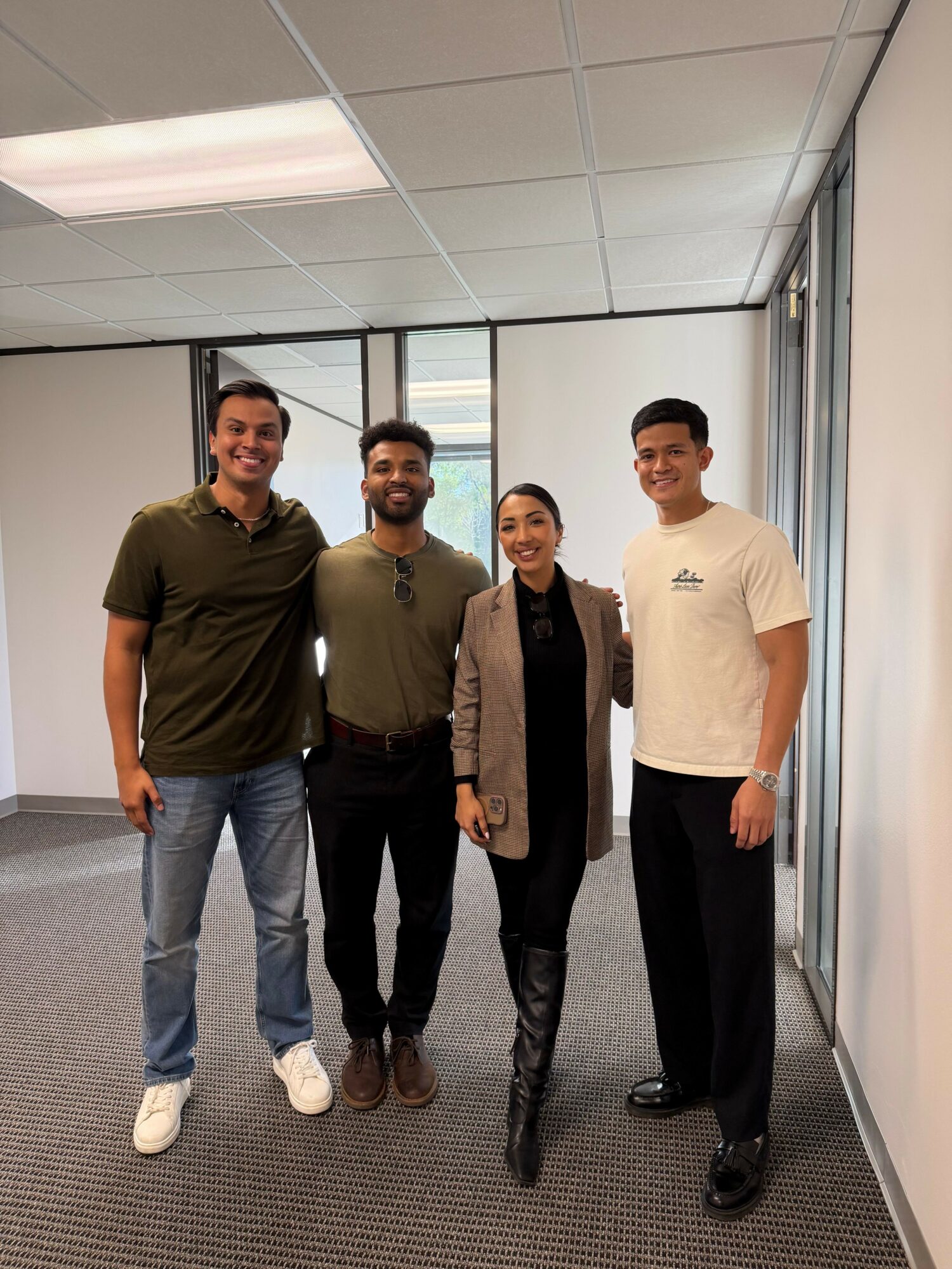 Four people standing together in an office, smiling, with windows and blinds behind them.