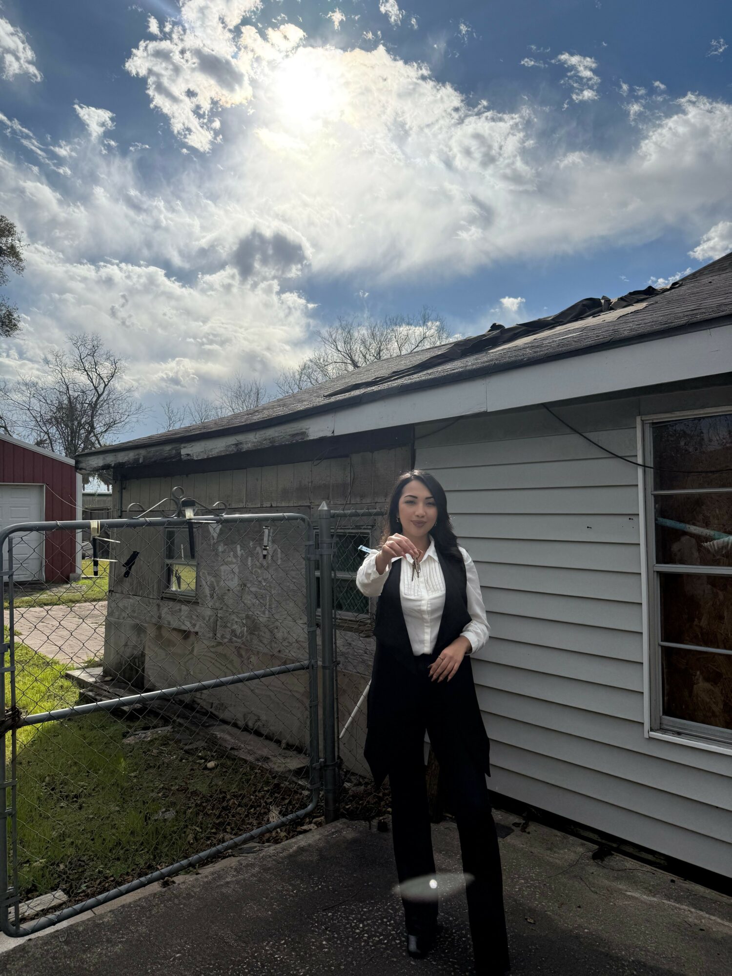 Woman standing outside near a house and fence, with cloudy sky above.