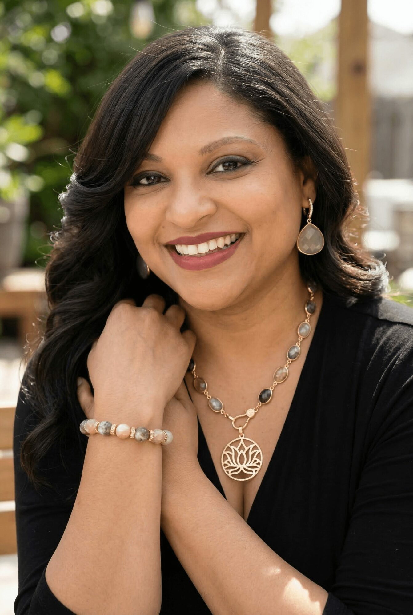 Woman with dark hair smiling, wearing earrings and a necklace, outdoors with greenery in background.