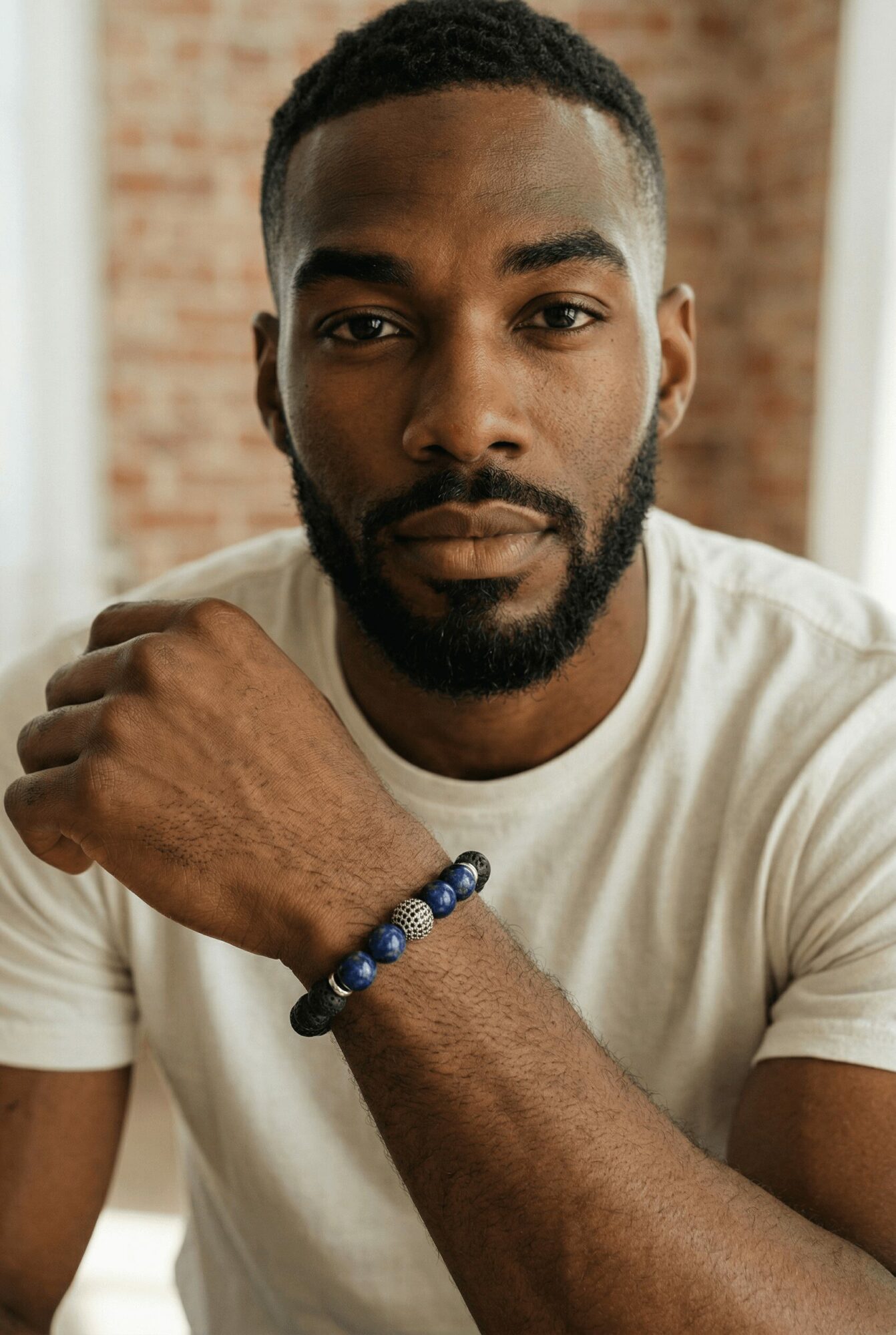 Man with short hair and beard wearing a white t-shirt, resting his arm on a surface, in front of a brick wall.