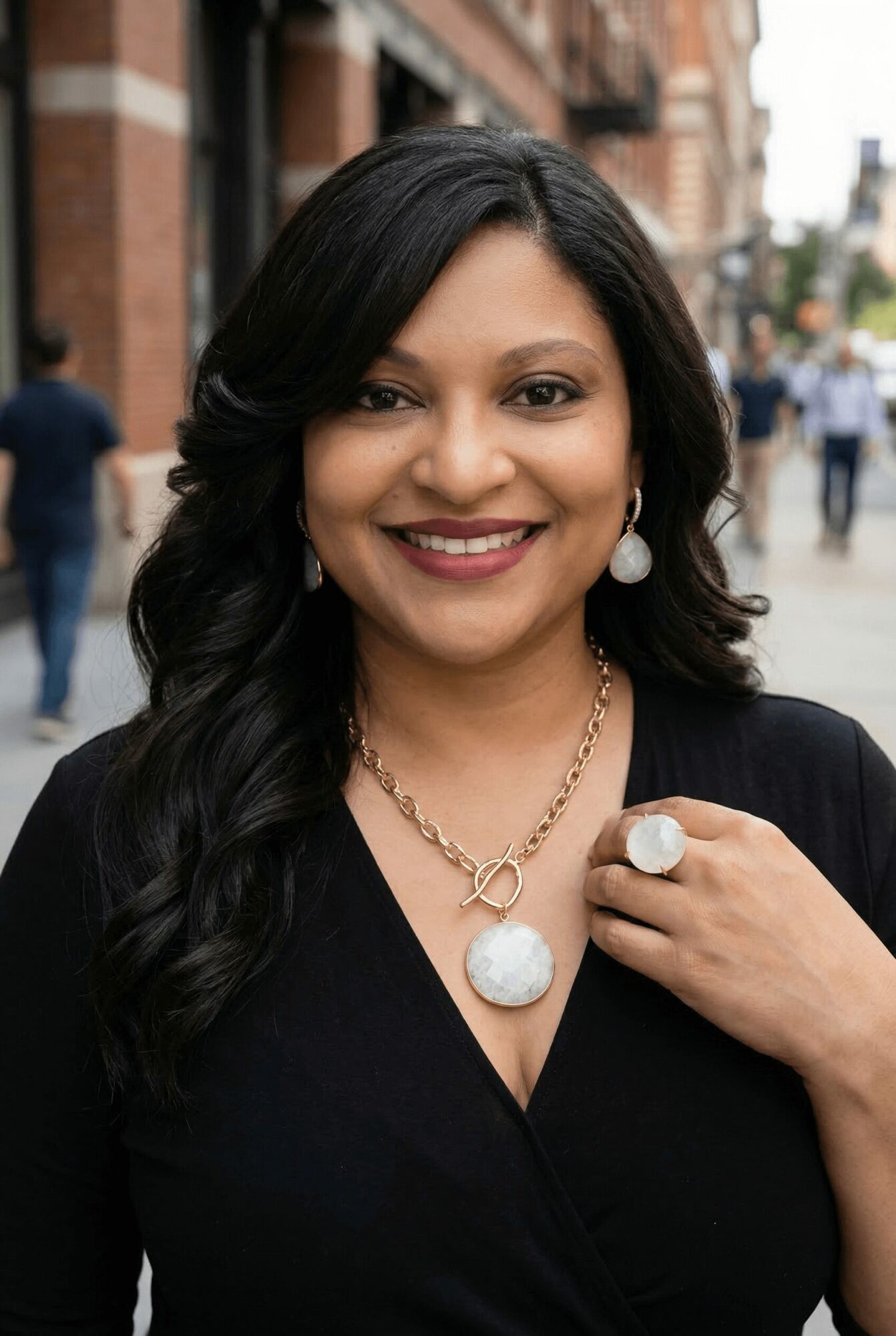 Woman with long dark hair smiling, wearing jewelry, outdoors in an urban setting.