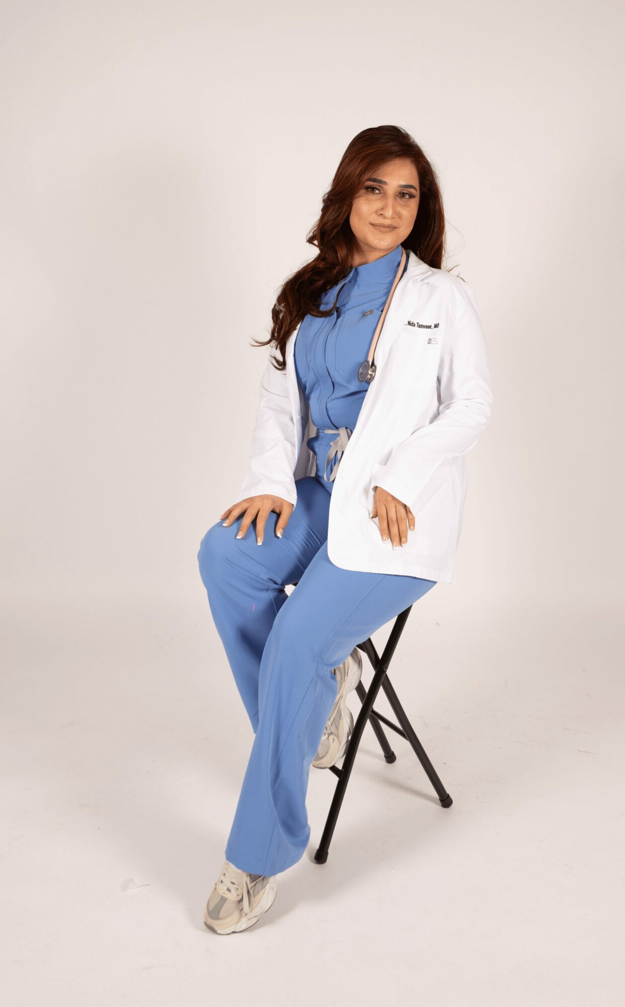 Woman in blue medical scrubs and white lab coat sitting on a black stool against a plain background.