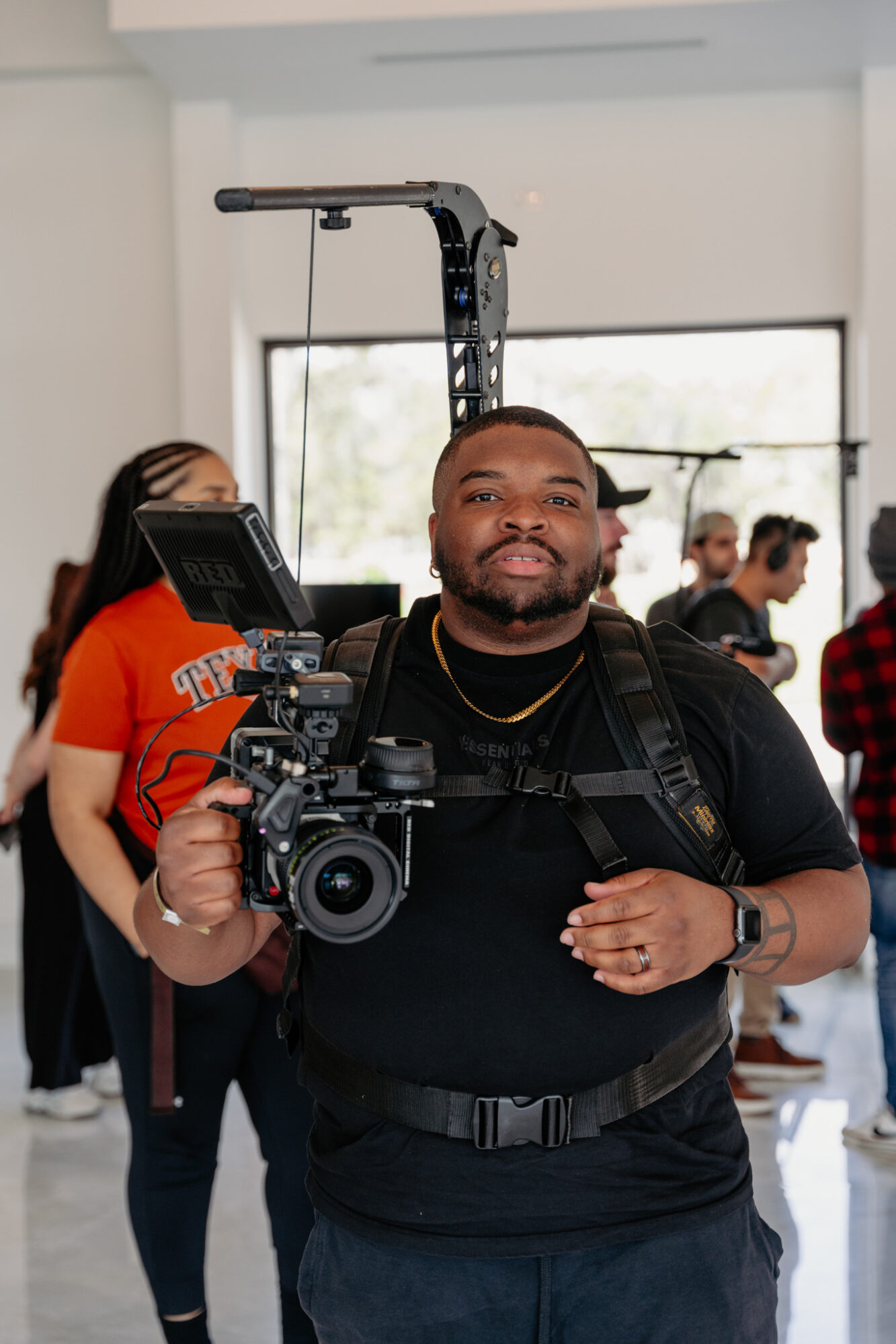 Man holding professional camera equipment indoors, with people in background, some wearing masks, and a window behind him.