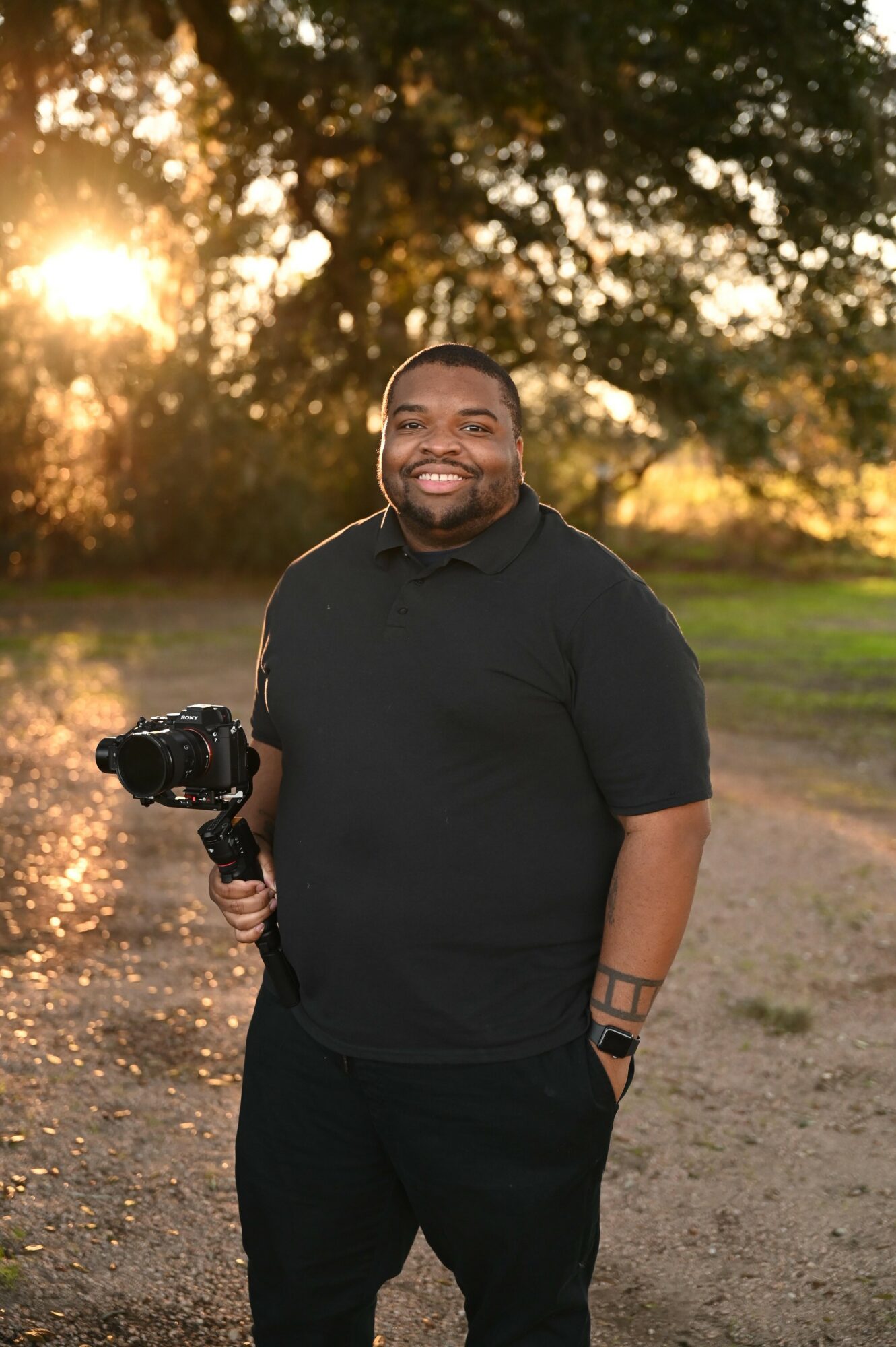 Man standing outdoors holding a camera, smiling, with trees and sunlight in the background.
