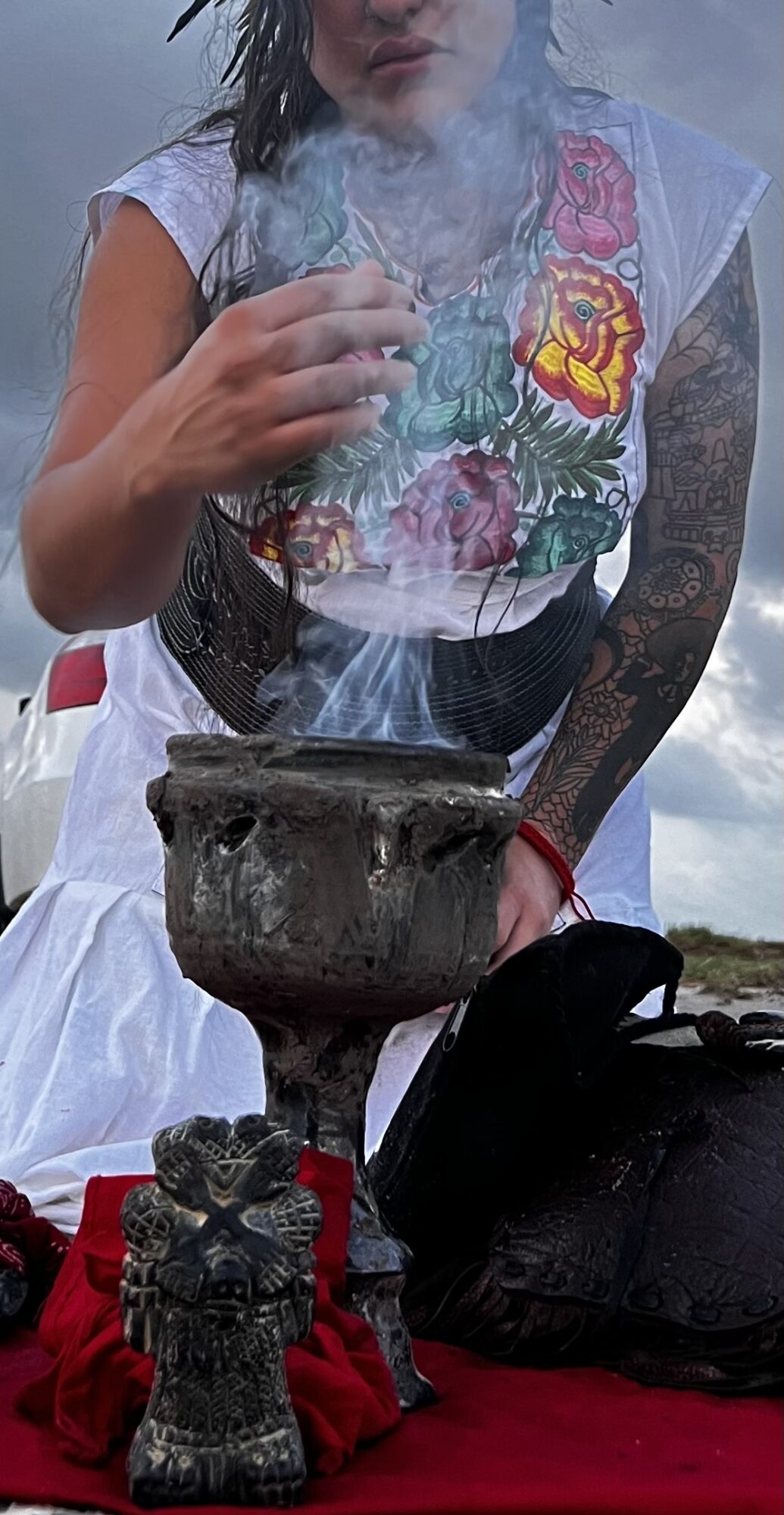 Person holding a smoking bowl with two small figurines in front, outdoors with cloudy sky background.
