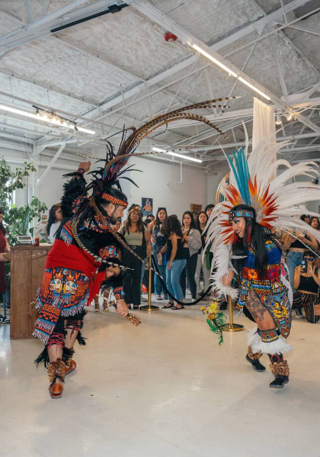 Two performers in colorful traditional costumes dance indoors with audience watching.