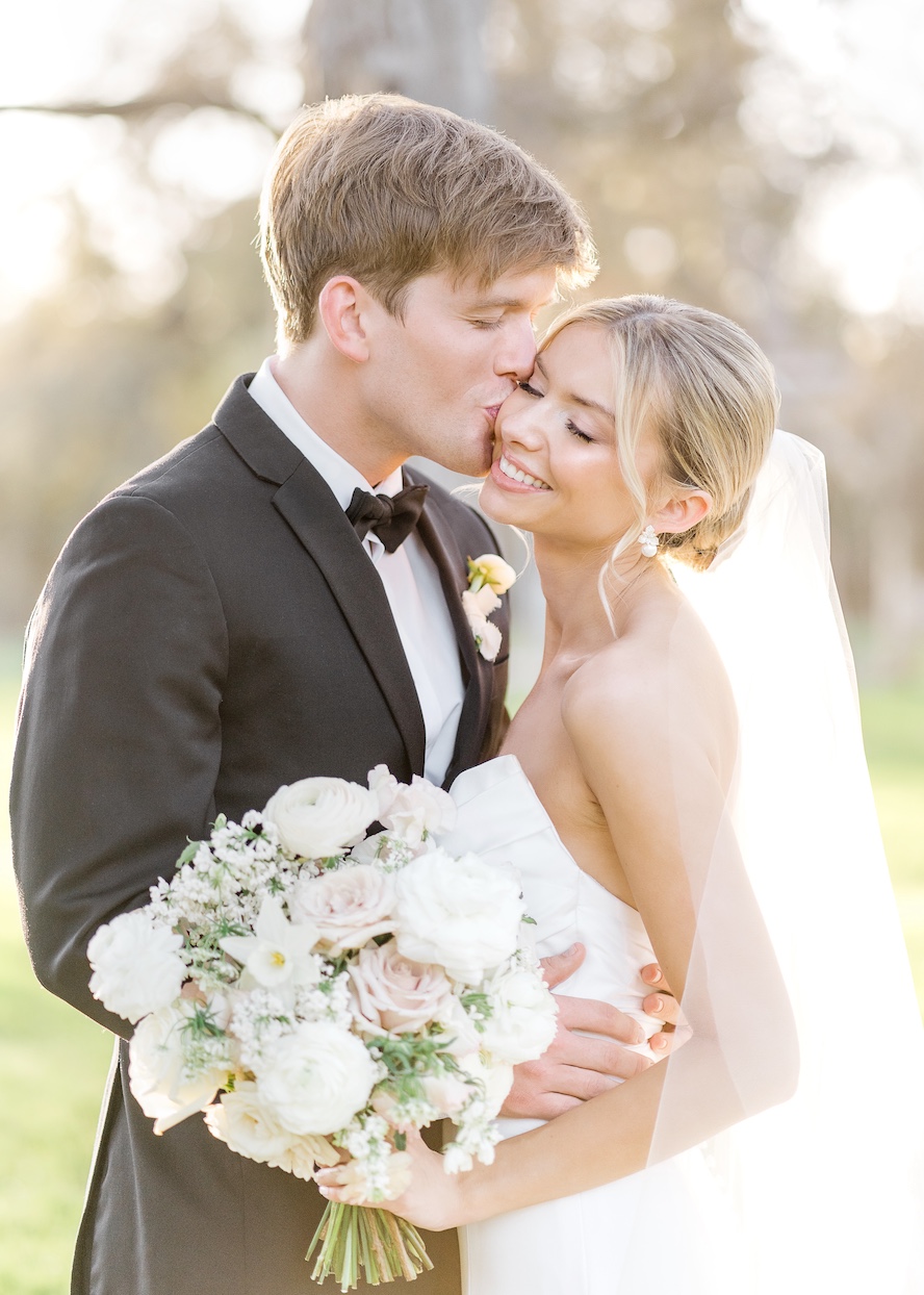 Bride and groom smiling outdoors, bride holding a bouquet, groom in tuxedo, bride in wedding dress with veil.