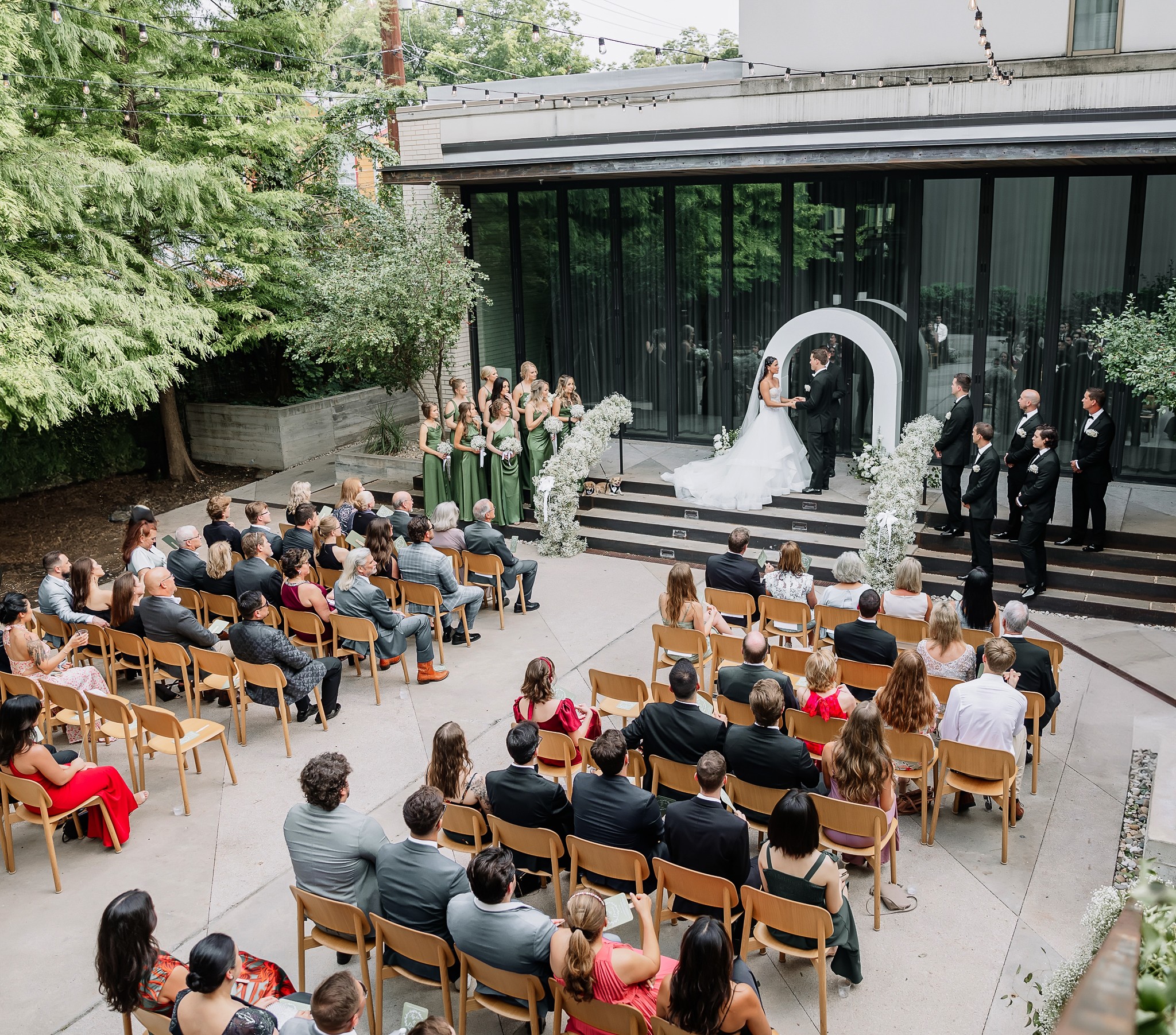 Wedding ceremony outdoors with guests seated, couple at altar, officiant, and wedding party, in front of modern building with glass windows.
