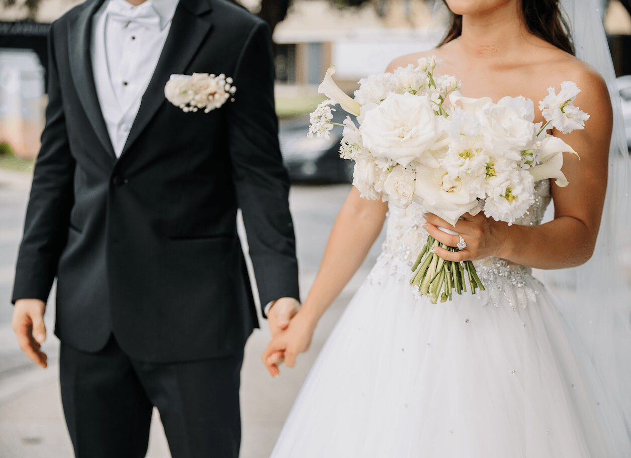 Bride holding a large bouquet of white flowers, wearing a white wedding dress. Groom in a black tuxedo with a white boutonniere.