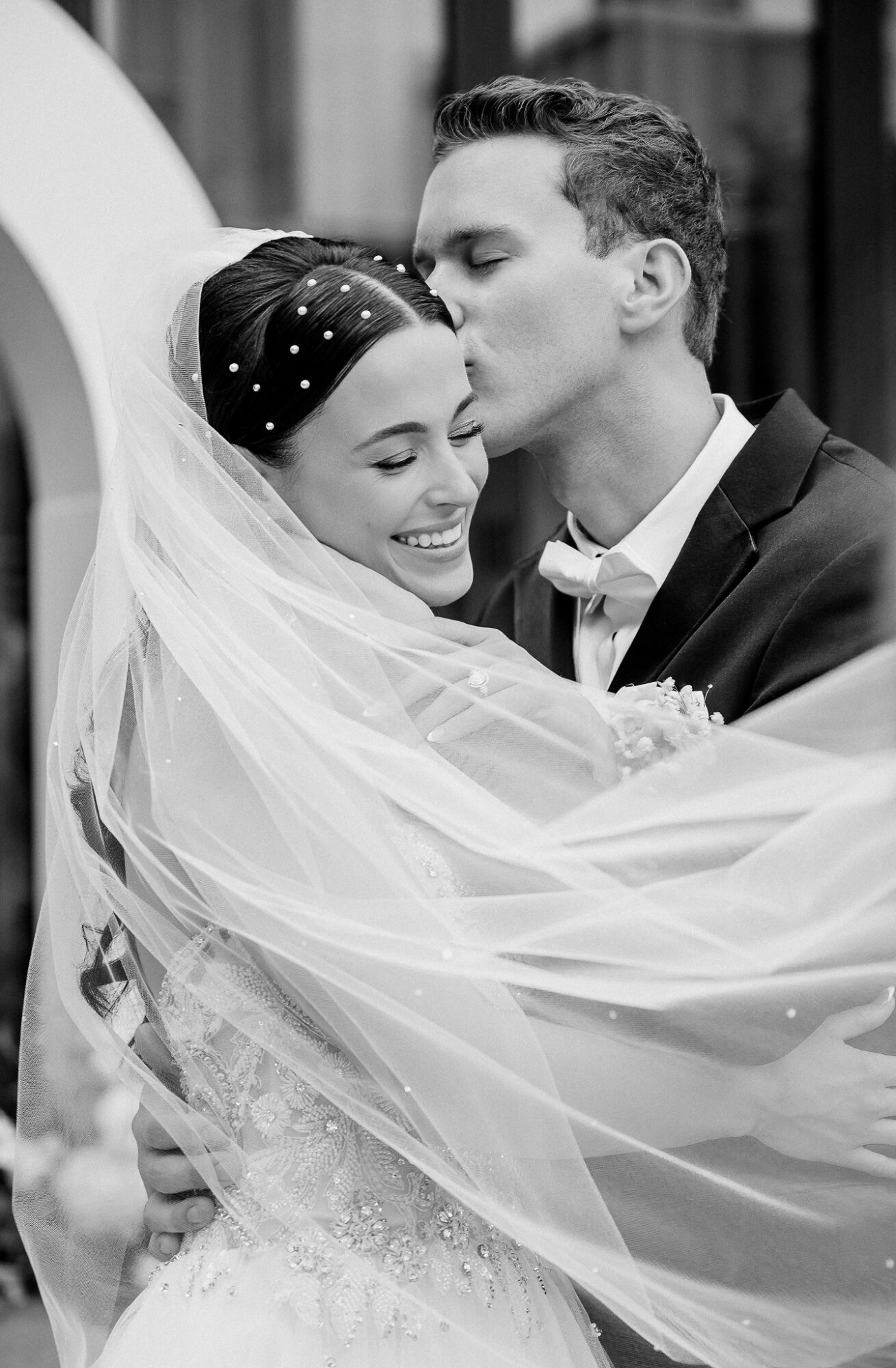 Bride and groom embrace, bride smiling, groom kissing her forehead, wedding attire, veil, outdoor setting.