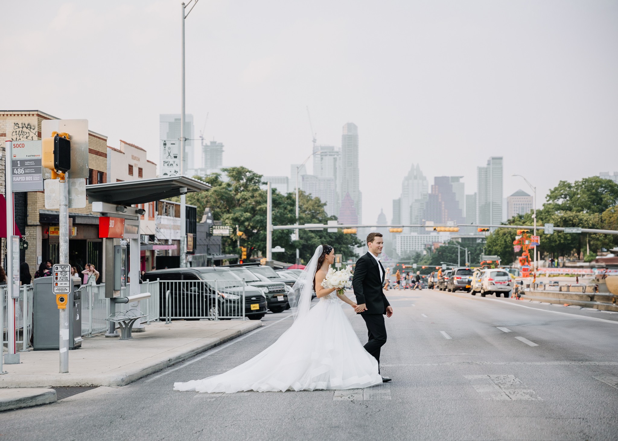 Bride and groom standing in the middle of a city street with buildings and traffic in the background.