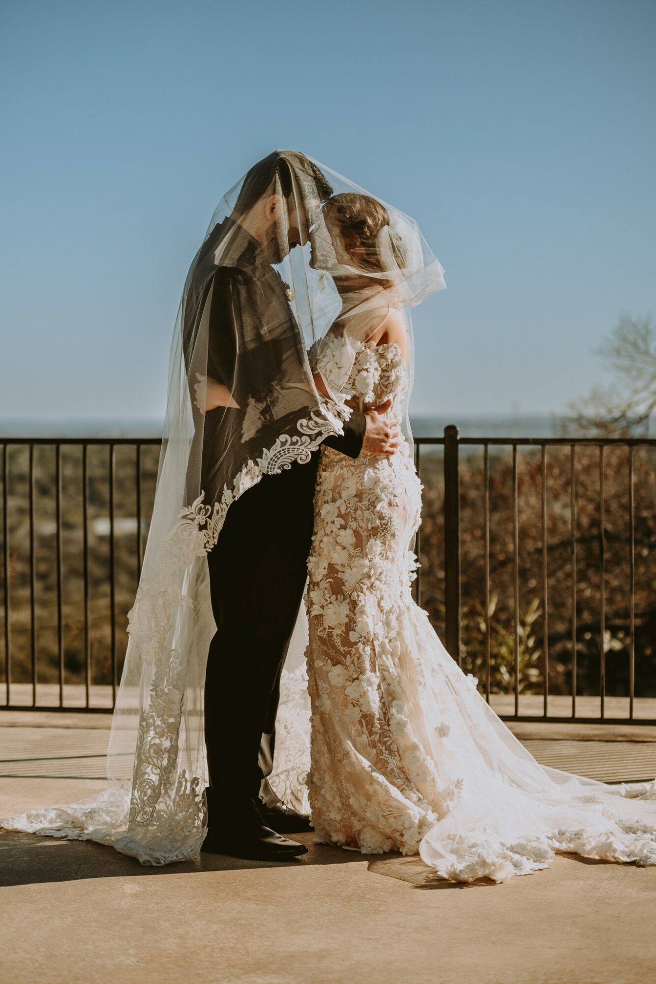 Bride and groom embrace outdoors, bride in lace wedding dress, groom in dark suit, veil covering both faces, railing behind them.