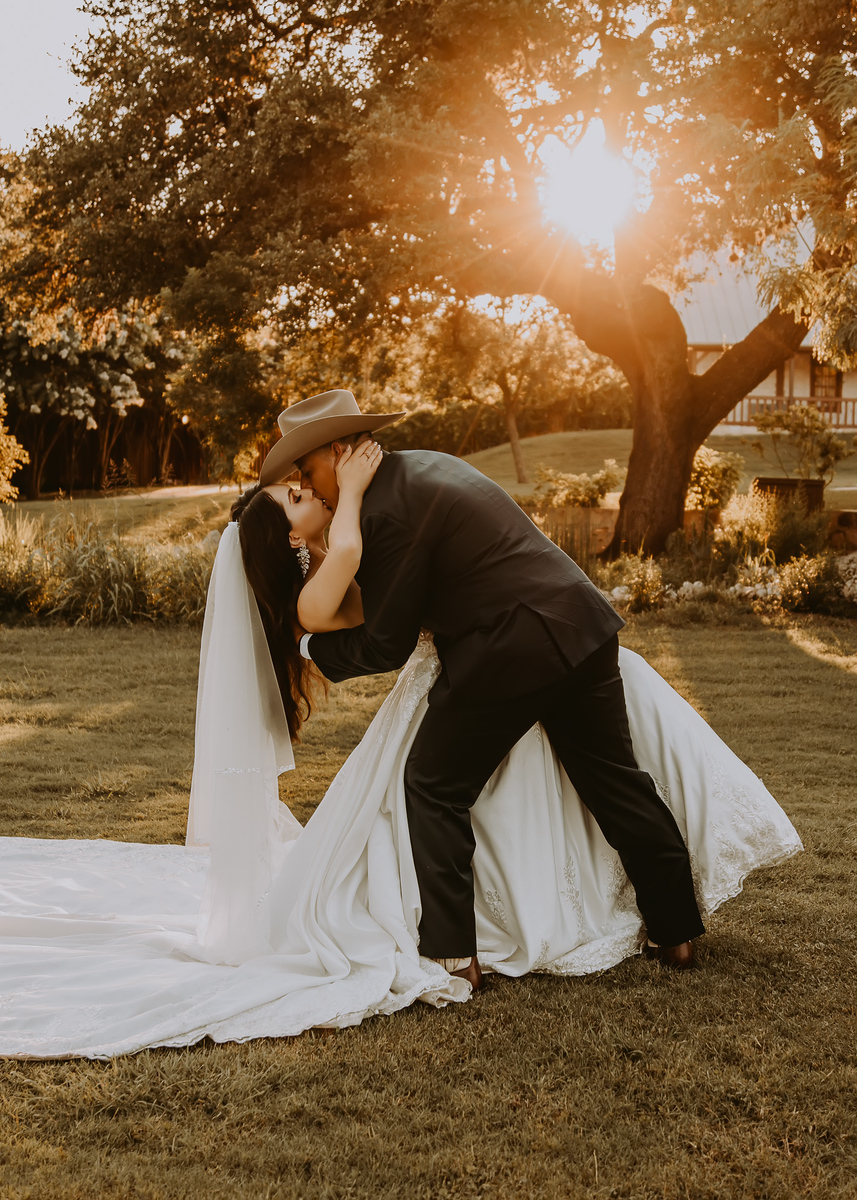 Couple kissing outdoors with sunlight filtering through trees, bride in white dress and veil, groom in dark suit, grassy area.