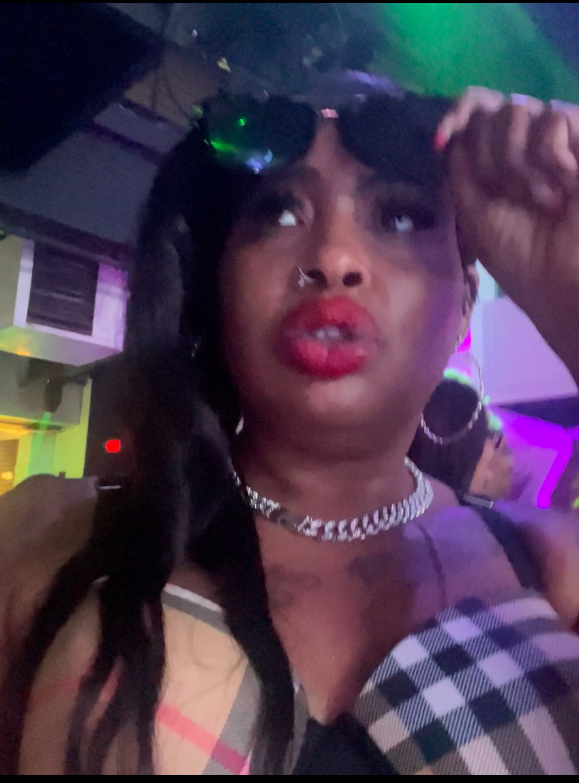 Woman with long dark hair, wearing hoop earrings, a necklace, and a checkered top, in a colorful indoor setting.