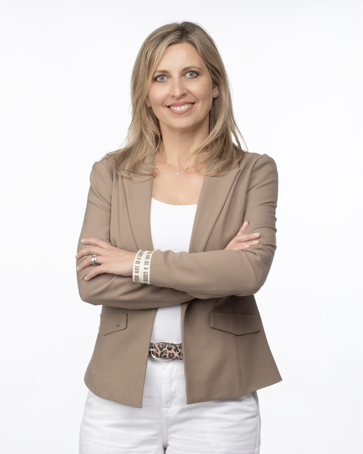 Woman with blonde hair smiling, arms crossed, wearing beige blazer and white pants, standing against a white background.