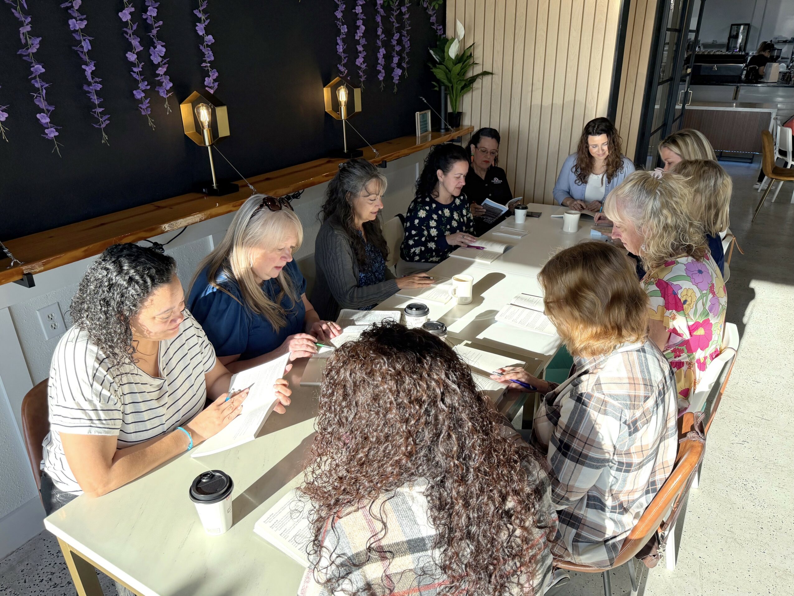 Group of women sitting around a long table in a room with purple flowers on the wall, some taking notes.