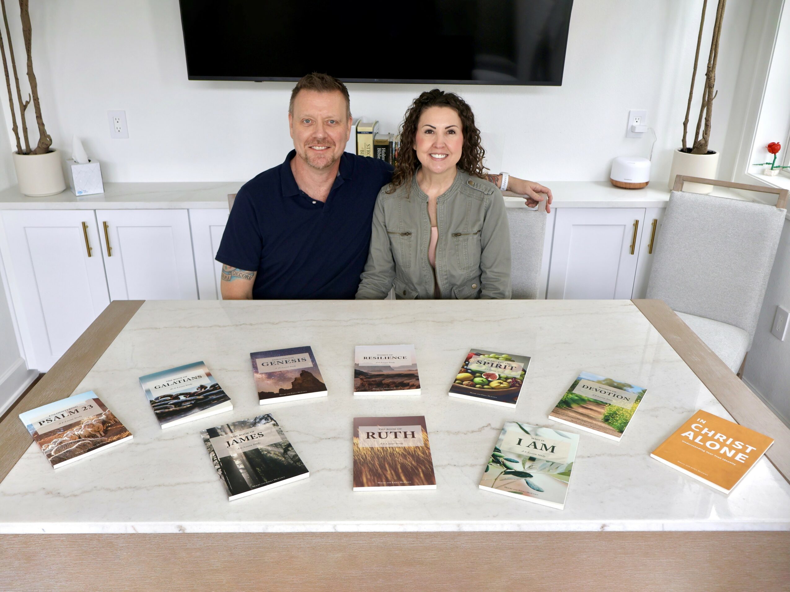 Two people sit at a table with books spread out, smiling in a bright room with white cabinets and a TV.