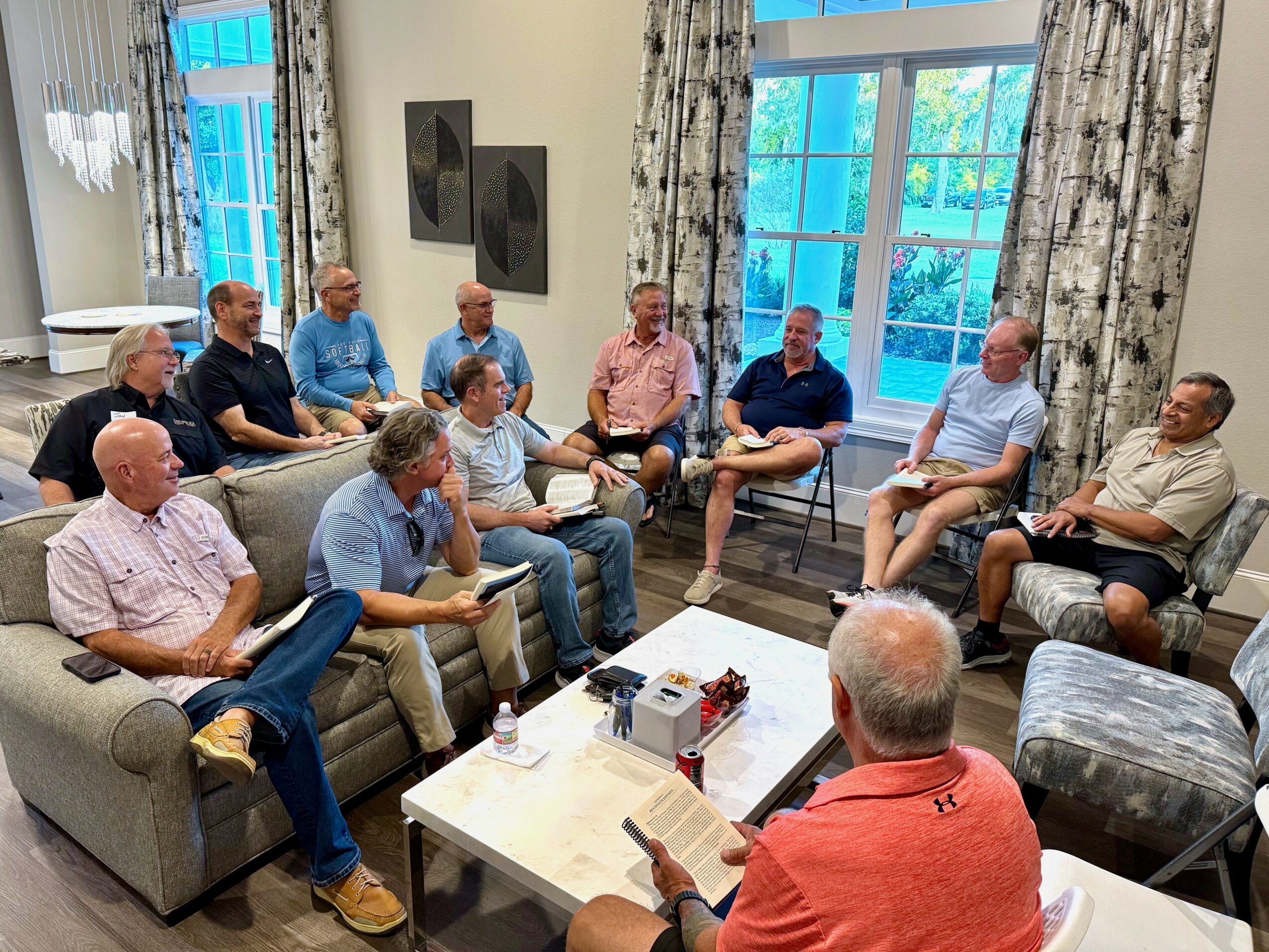 Group of people sitting in a living room, engaged in a discussion, with large windows and curtains in the background.