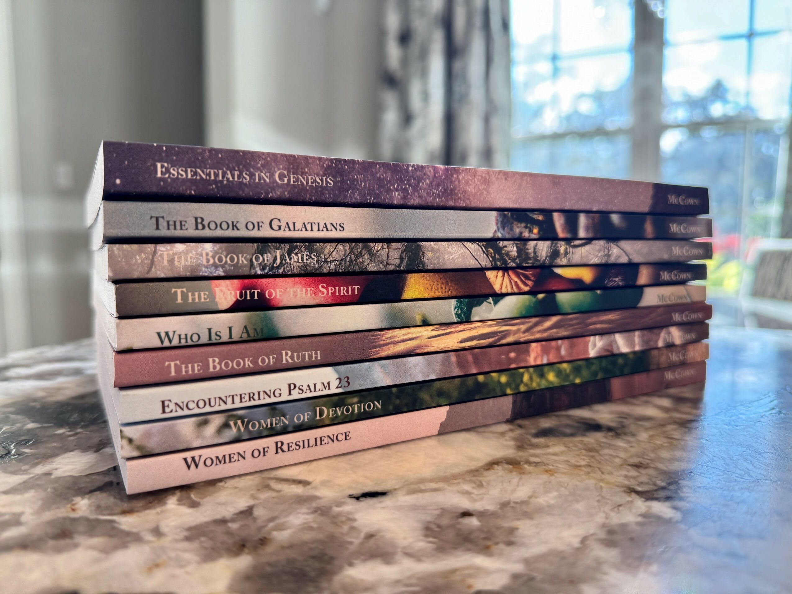 Stack of eight books with colorful covers on a marble surface, sunlight coming through window in background.