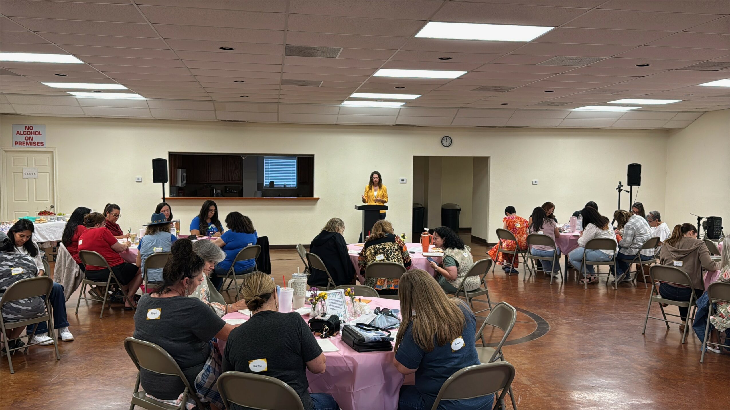 People seated at round tables in a large room, listening to a speaker at a podium, with a screen behind.