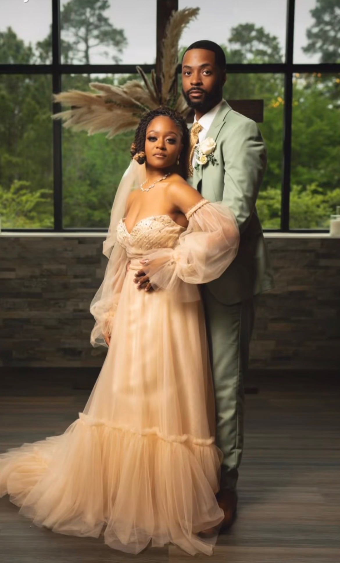 A couple dressed in formal attire standing indoors with large windows and trees outside.