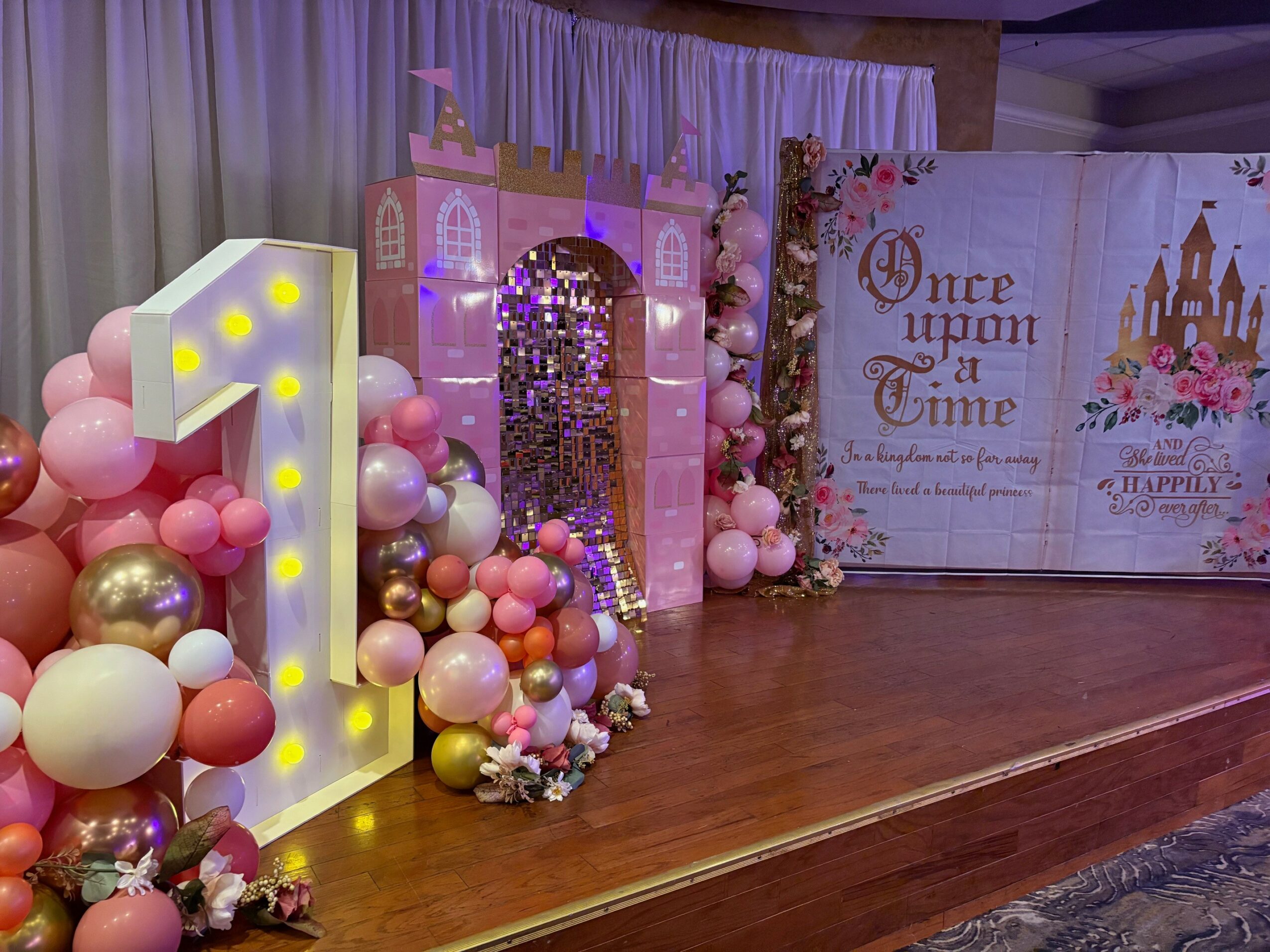 Decorative birthday setup with a large illuminated number one, balloons, a castle backdrop, and a banner reading 'Once upon a Time'.