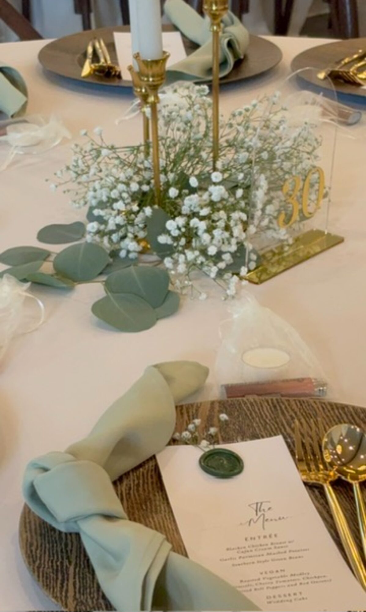 Table centerpiece with white flowers, eucalyptus leaves, gold candle holders, and a menu card on a table setting.
