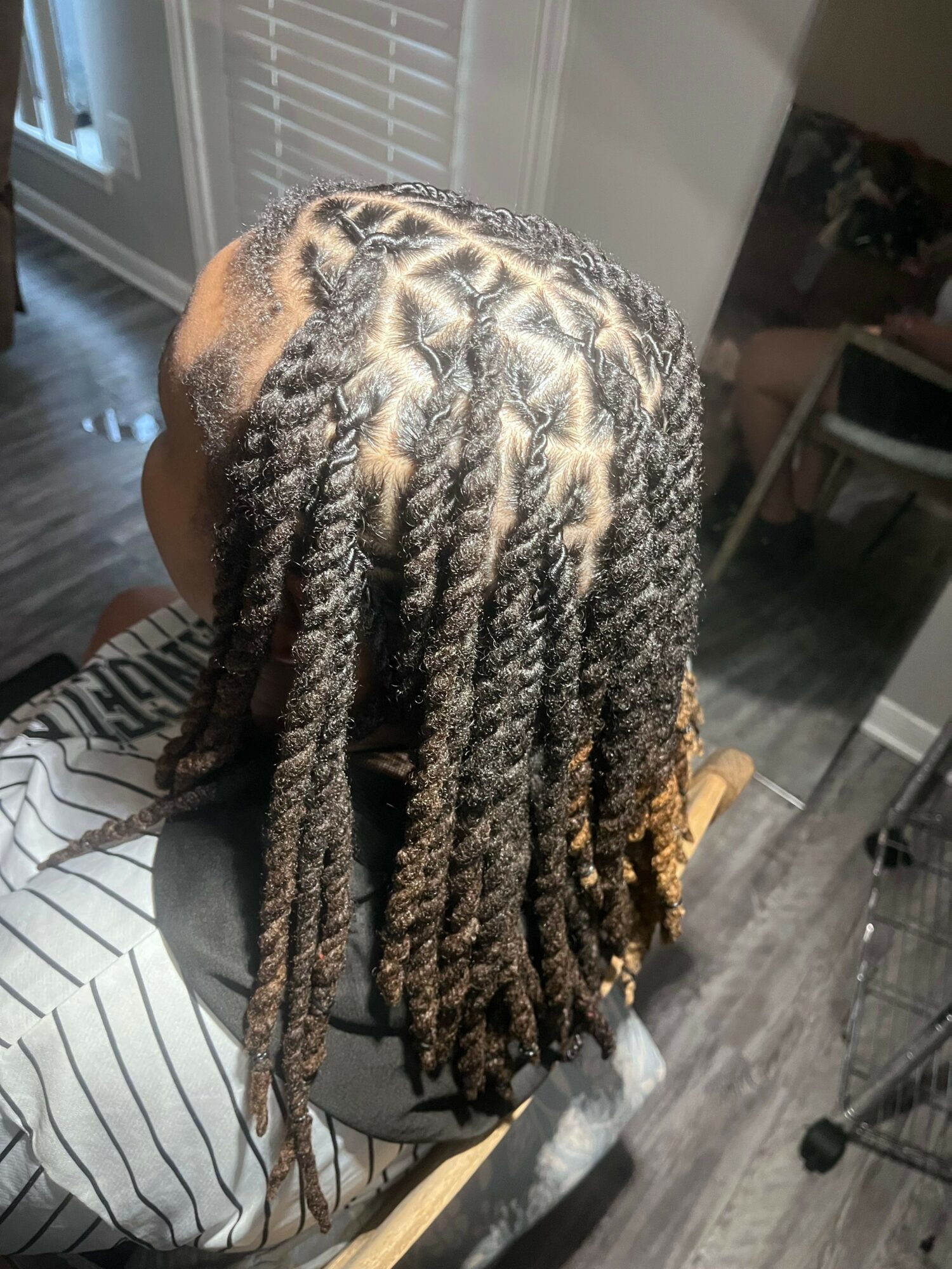 Person with neatly styled dreadlocks, viewed from the back, sitting indoors near a window and furniture.