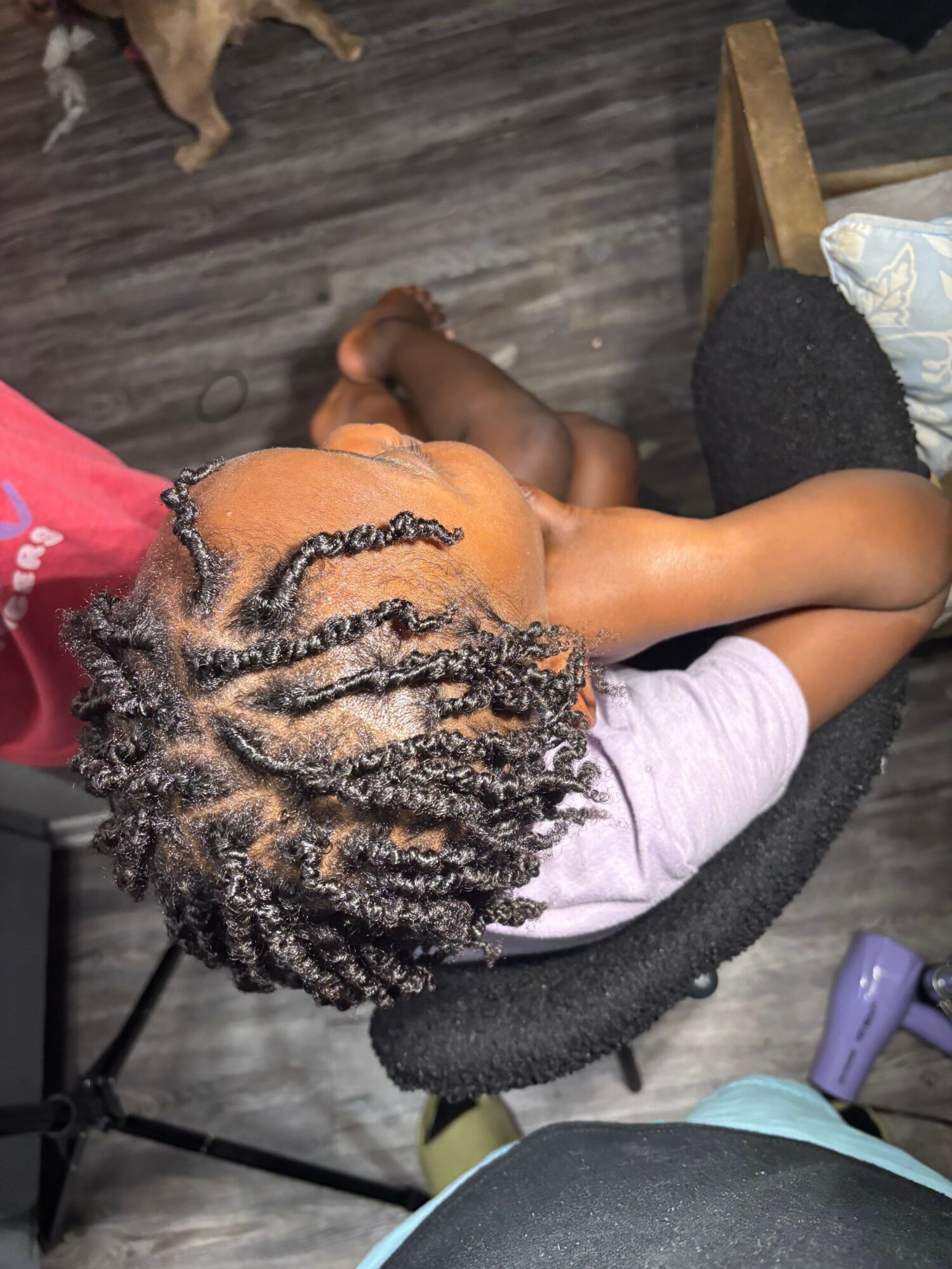 Child with styled hair sitting on a black chair, wearing a light purple shirt, with a wooden floor background.