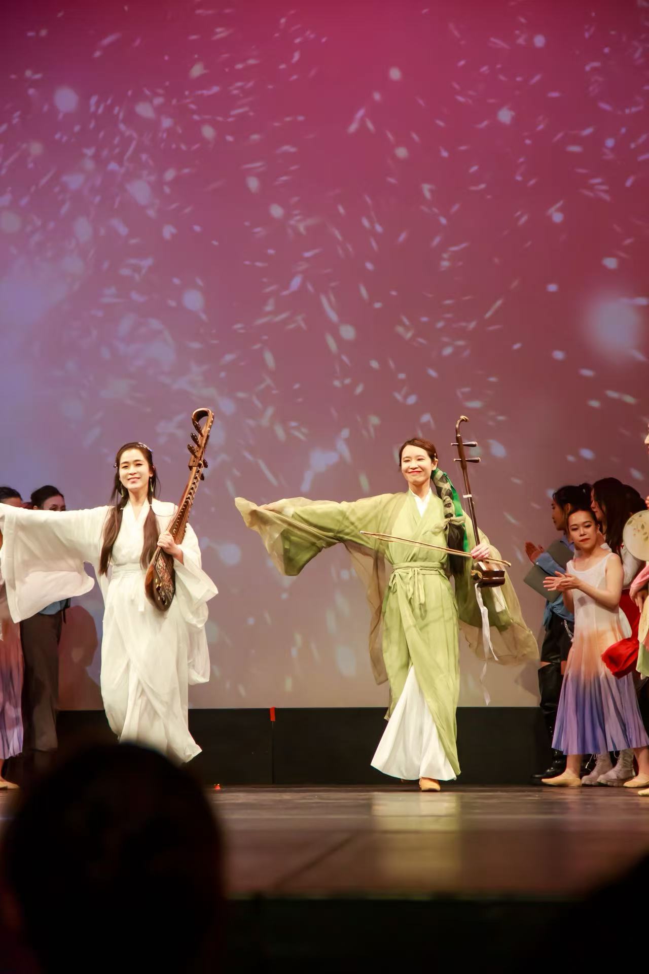Group of performers on stage, two women in traditional costumes holding musical instruments, others in colorful attire, stage background pink.