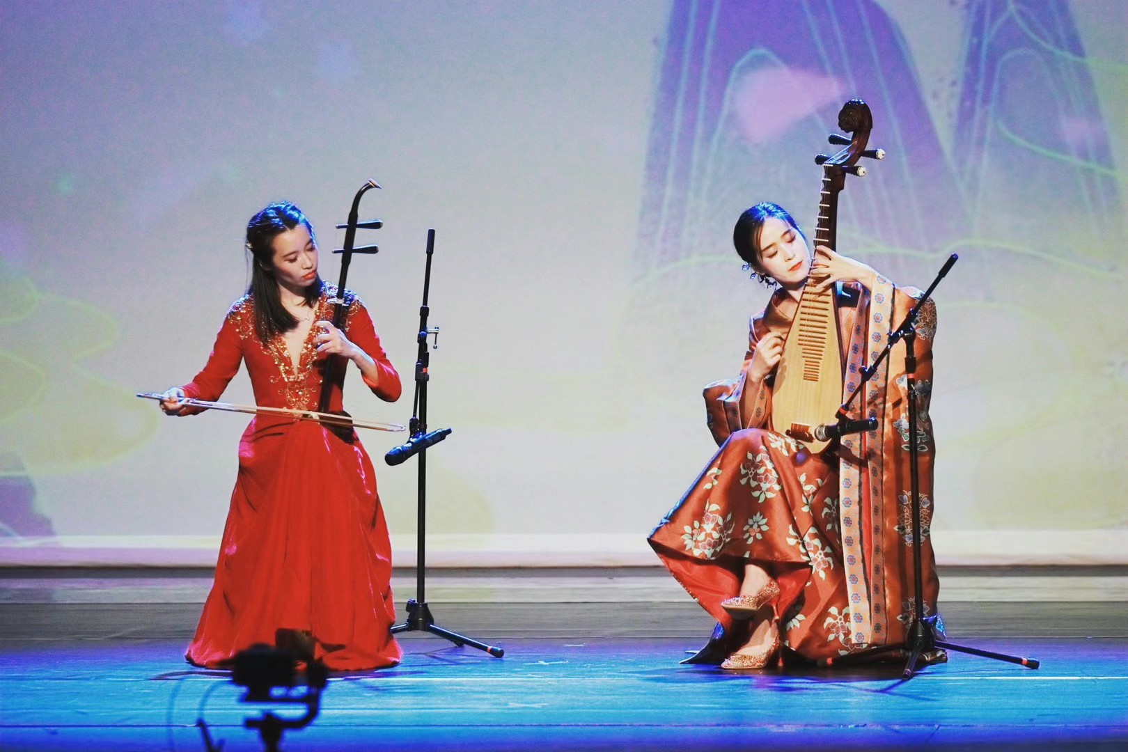 Two women in traditional dresses perform on stage with musical instruments, one singing and the other playing a string instrument.